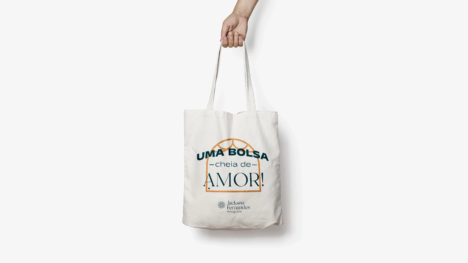 Mão segurando uma ecobag de pano na cor crua, estampada com a frase 'UMA BOLSA cheia de AMOR!' dentro de um arco laranja, seguida pelo logotipo Jackson Fernandes Fotografia.