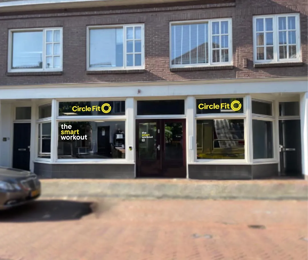 A Circle Fit hub with large windows displaying yellow signs, located on a brick street with a parked car nearby.