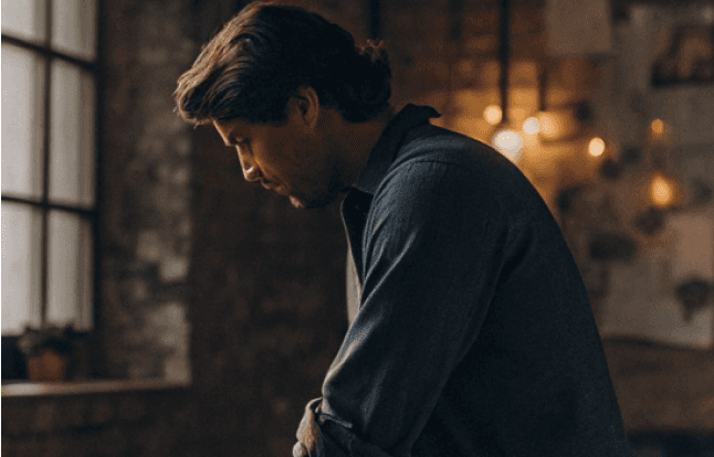 Man with dark hair looking down in deep thought. He wears a button-down shirt, near a window.