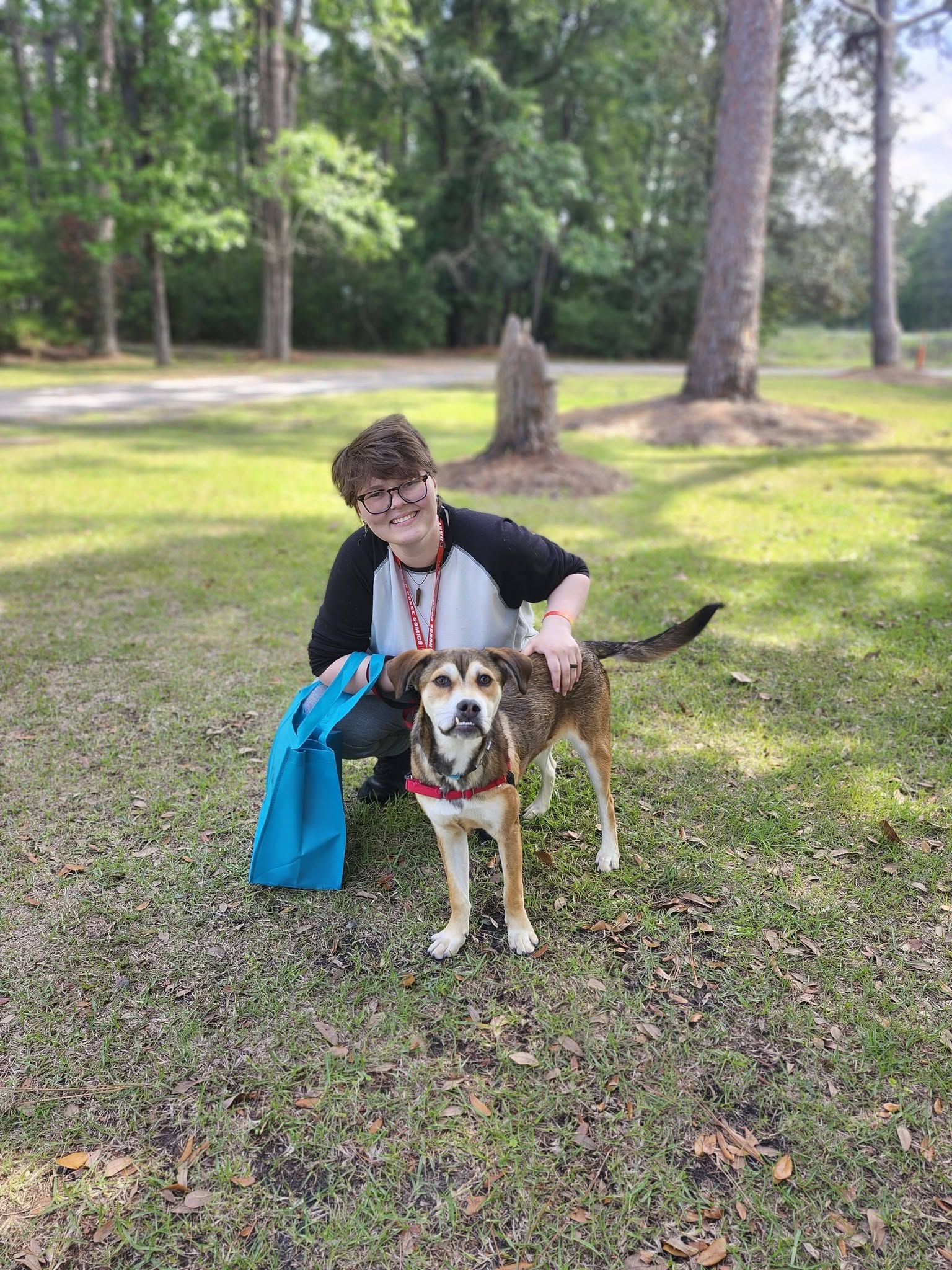 Un adoptante feliz arrodillado junto a un perro de rescate marrón y blanco recién adoptado en un parque con césped.