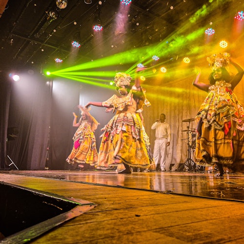 身穿色彩鲜艳服装的表演者在舞台上充满活力地舞动，舞台上洒满了明亮的黄色和绿色灯光。