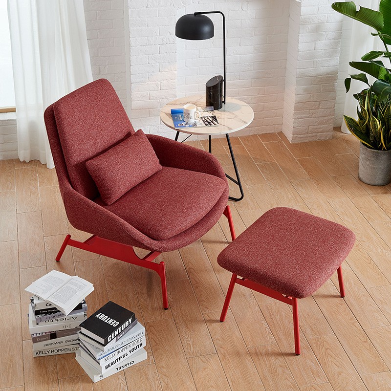 High-angle view of a red mid-century modern lounge chair and ottoman set in a cozy reading nook, featuring a stack of books, marble side table with a black lamp, and a white brick fireplace backdrop.