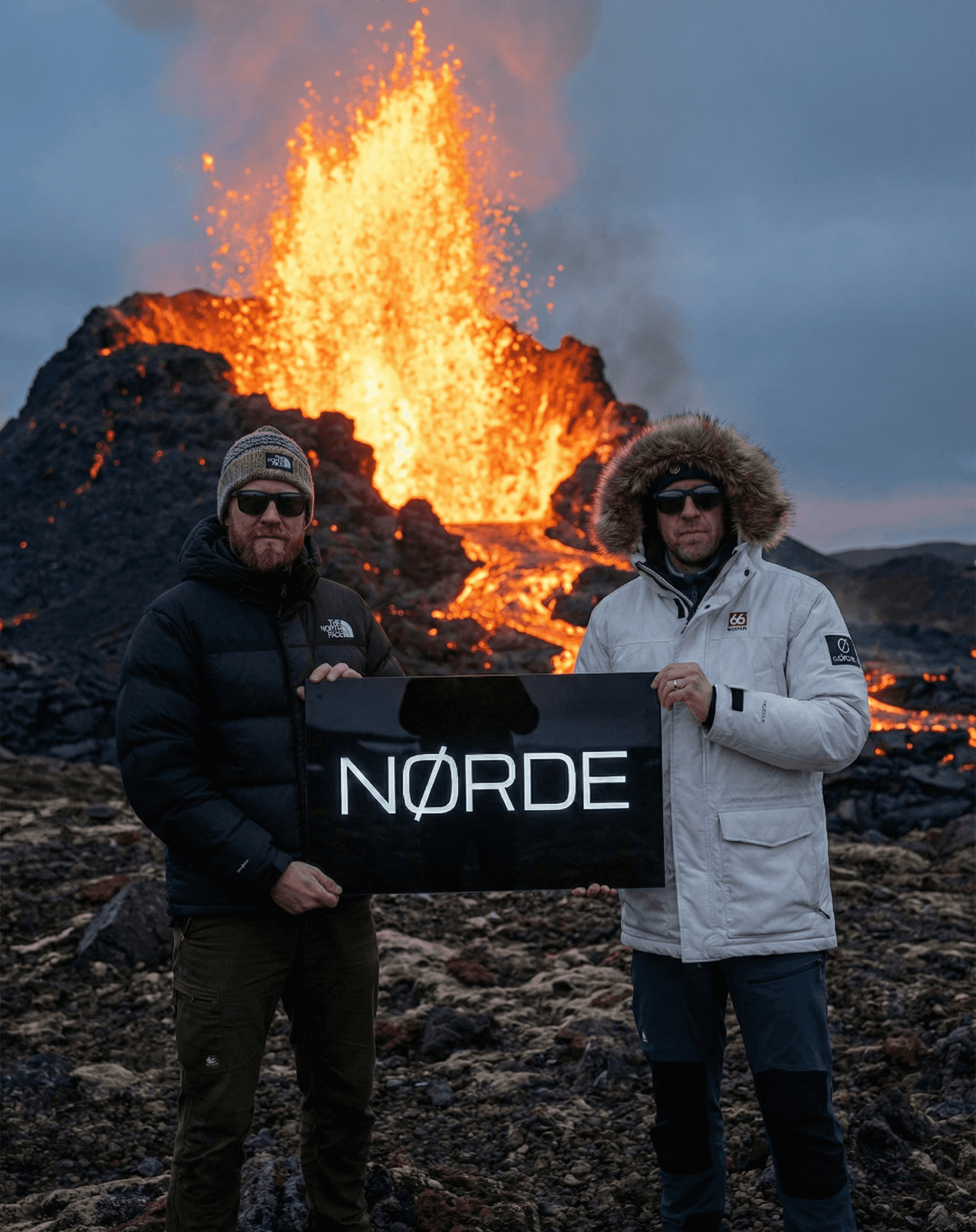 Two men in winter jackets and sunglasses standing in front of an active volcanic eruption with flowing lava, holding a black sign together.