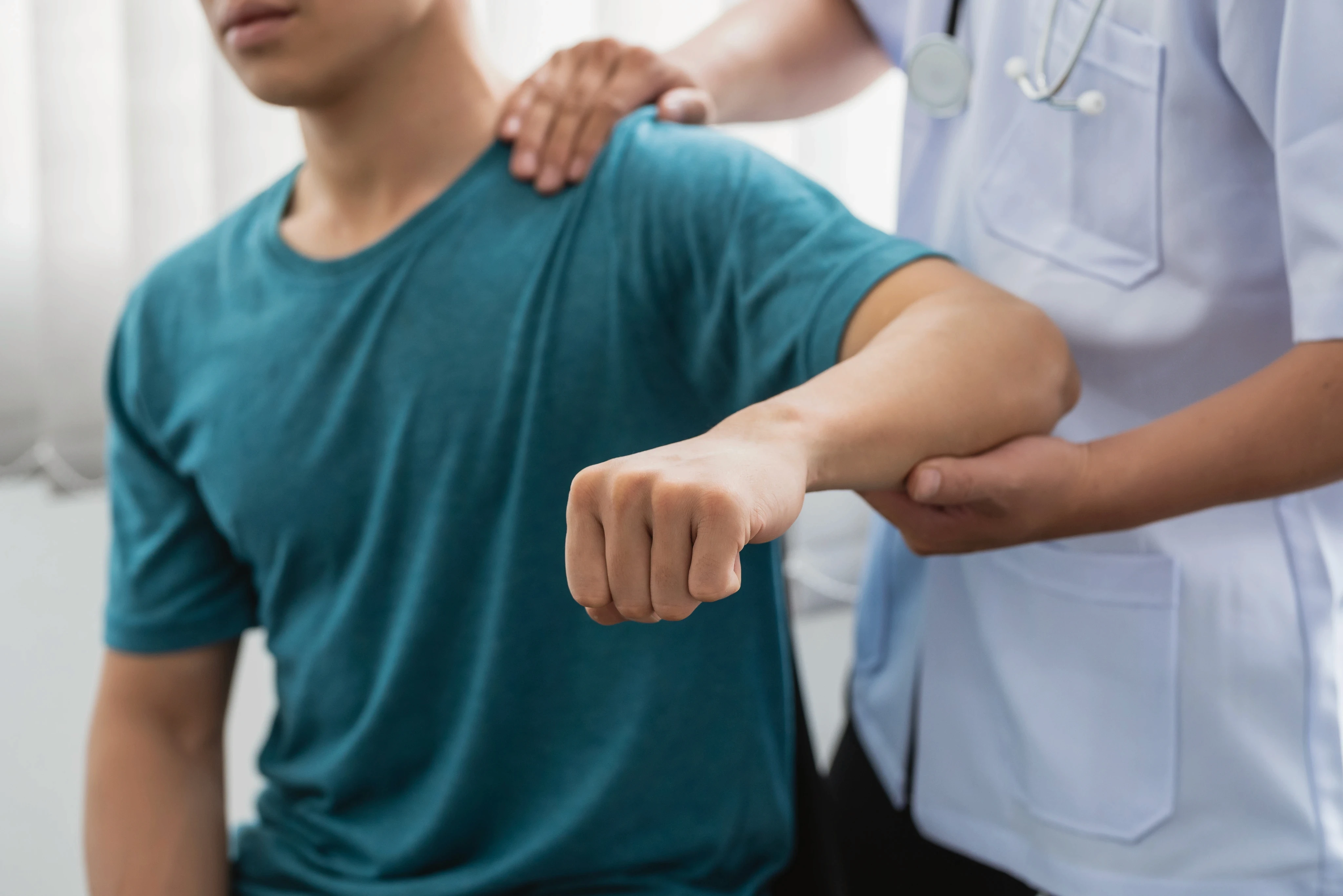 A person in a teal shirt sits while a healthcare professional checks their arm movement, conveying a sense of medical care and physical assessment.