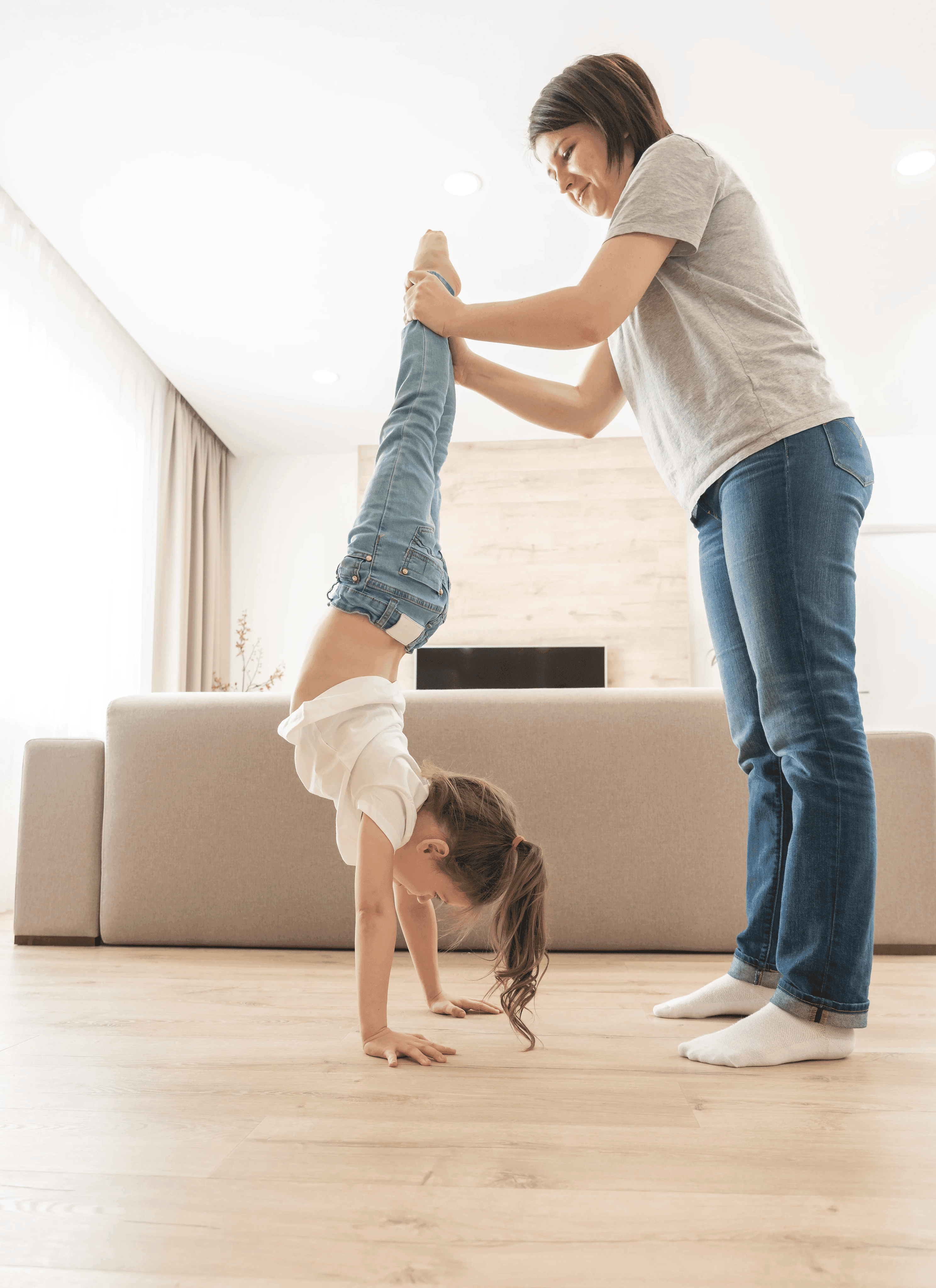 A mother supports her young daughter as she practices a handstand on LUXO’s hybrid flooring, showcasing the floor’s soft feel, slip-resistant surface, and durability. The bright living room and natural wood tones highlight how LUXO Floors create a safe, comfortable space where families can play, grow, and enjoy everyday moments at home.