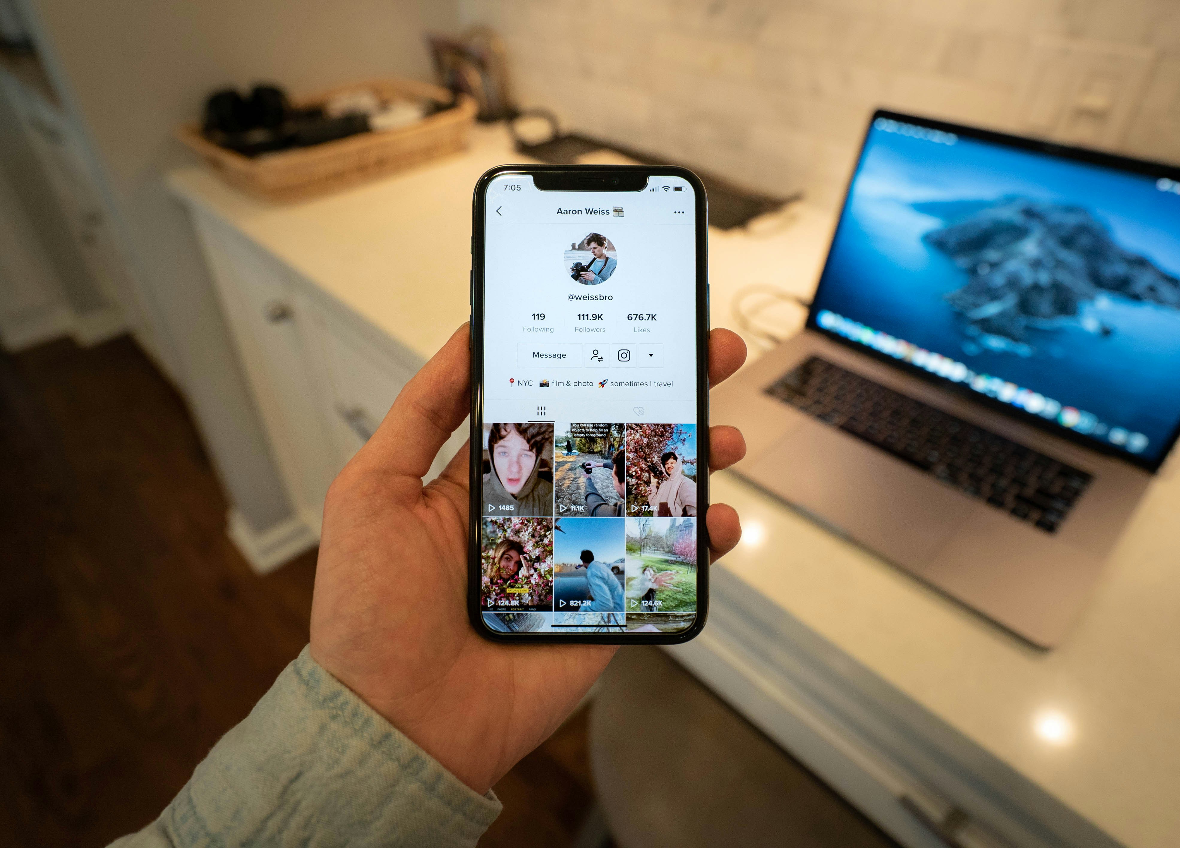 image of a person holding their phone while looking at a tiktok grid. Laptop in the background on desk