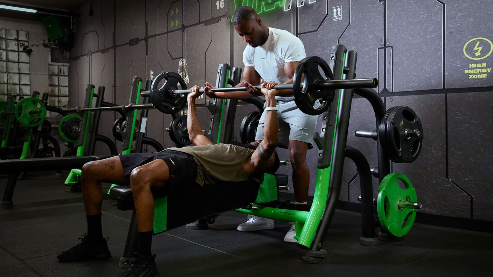 Zwei Männer im Fitnessstudio, einer führt Bankdrücken aus, während der andere als Spotter unterstützt.