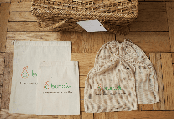 Three off-white fabric bags with green and orange logos are arranged on a wooden floor. The largest bag is positioned to the left, with two smaller bags to its right. All three bags display “From Mother” and the word “bundle” beneath their respective logos. A woven basket with a white tag rests on the wooden floor above the bags.