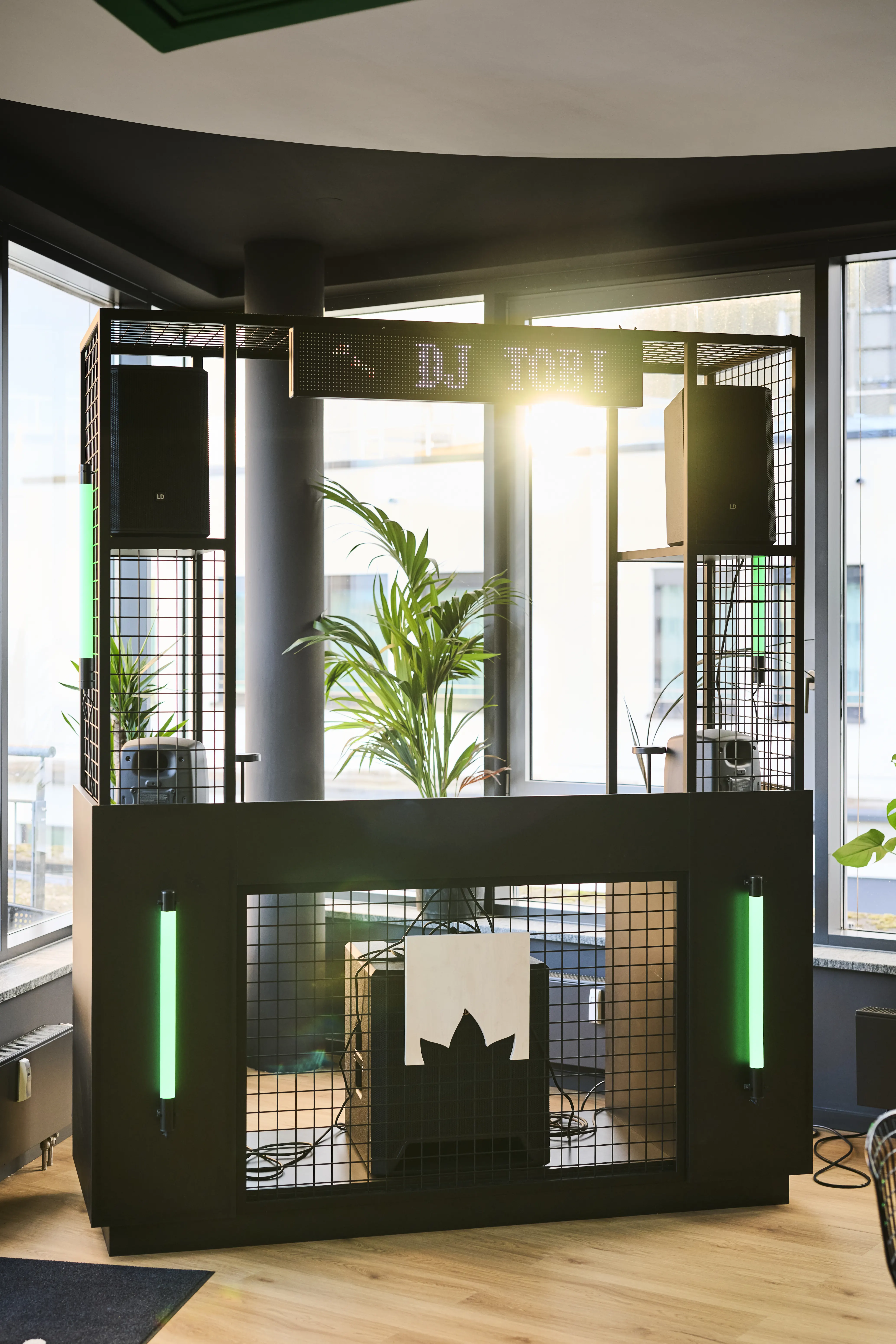 Custom DJ booth at BLOOMWELL made of black steel mesh with green neon tubes, mounted speakers, a DJ TOBI sign, a palm plant inside and a leaf logo panel.