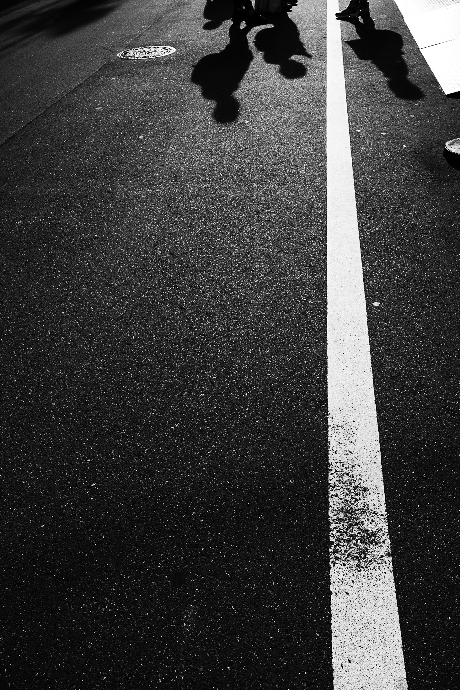 Black and white fine art photograph of human shadows crossing an urban street with strong sunlight and graphic asphalt textures