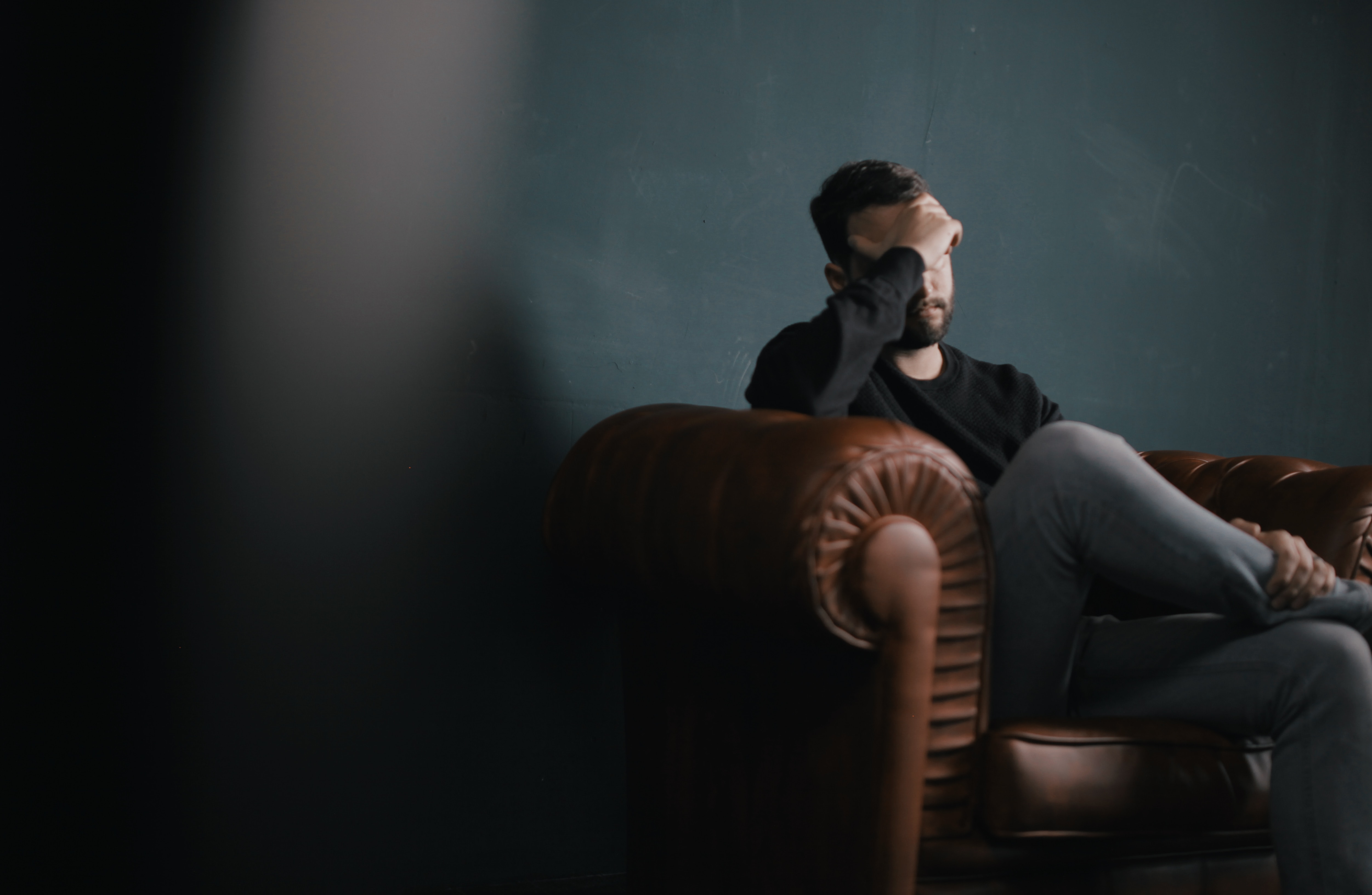 A man sat on a couch in a dark room, with his hand over his face 