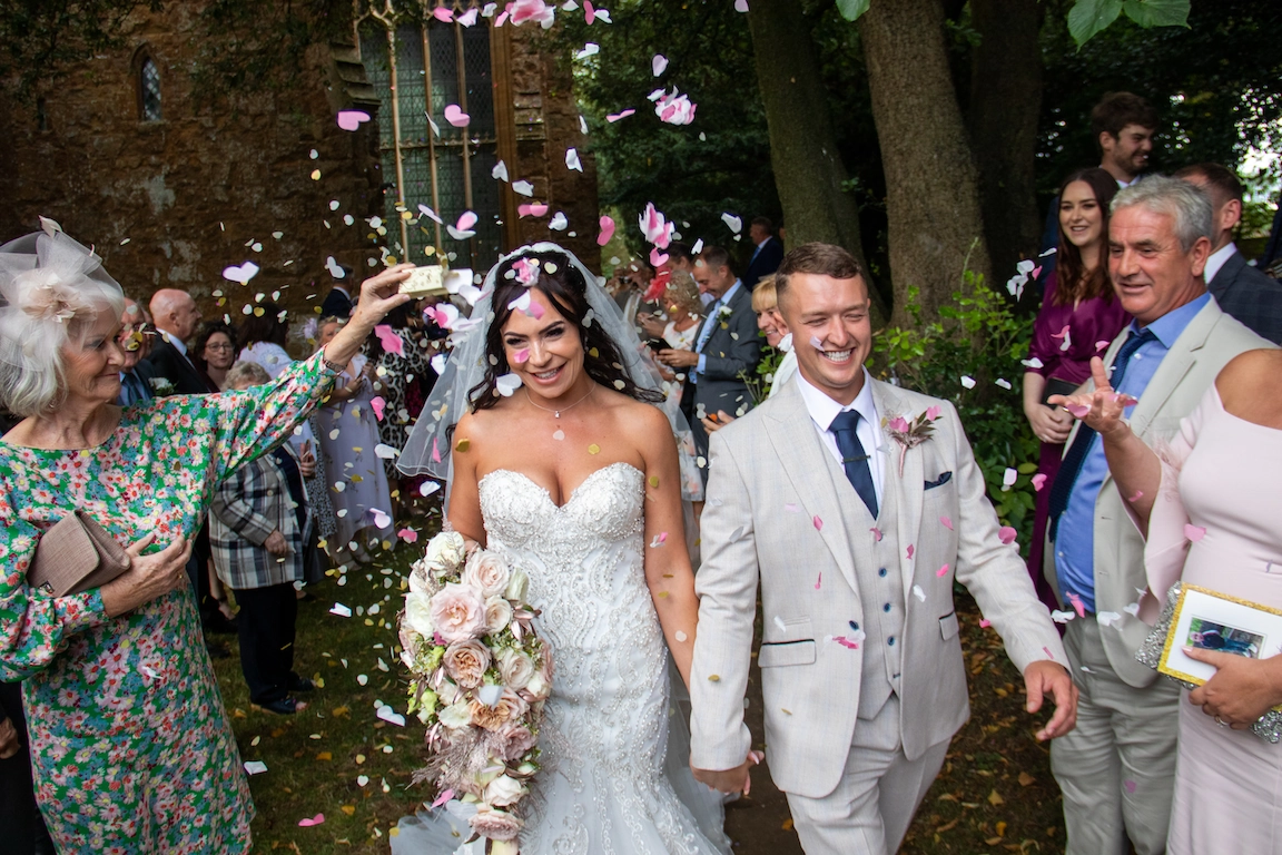 Bride and groom walking through confetti outside Bloxham Church after their wedding ceremony