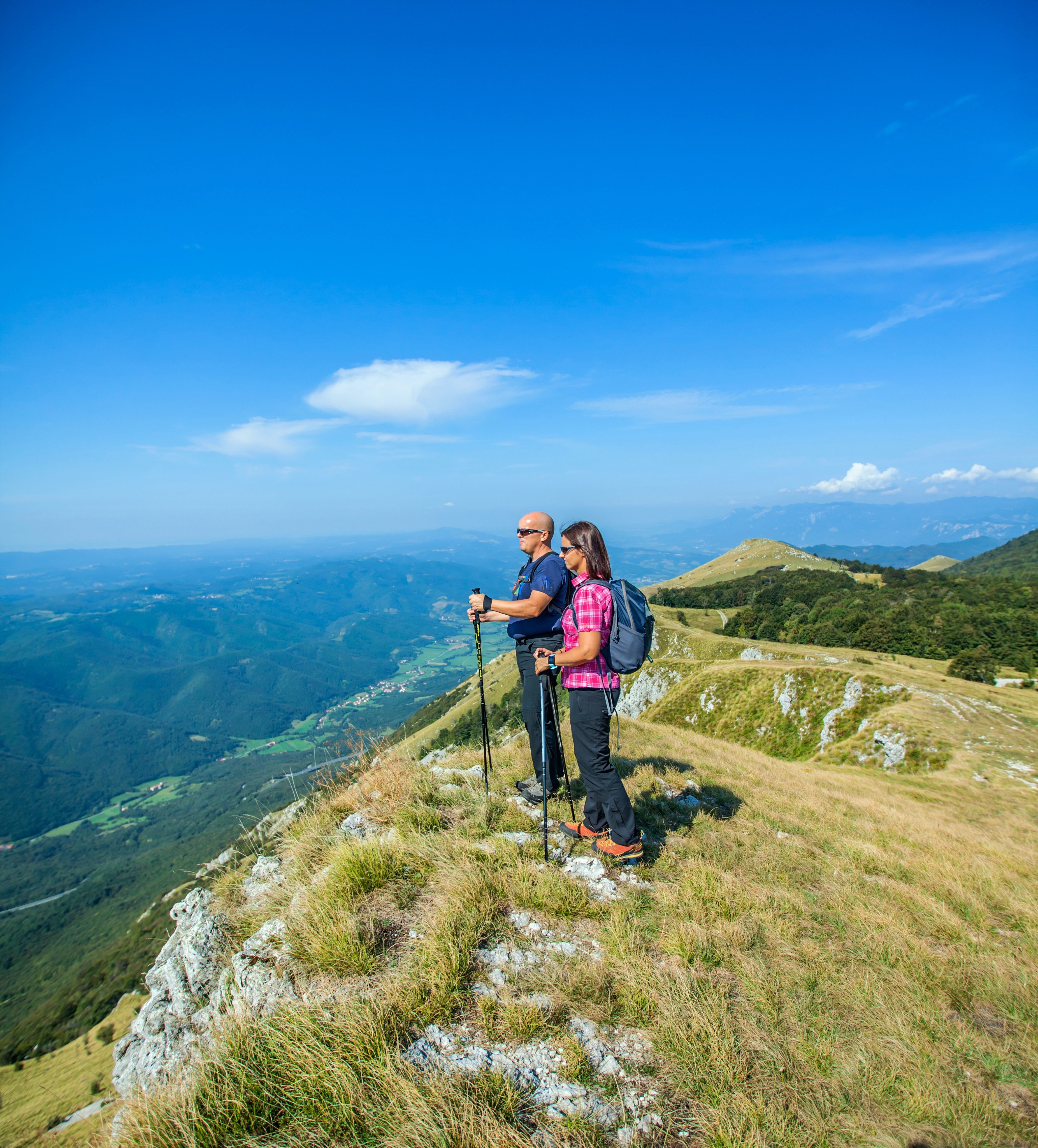 Two hikers standing on a grassy mountain ridge overlooking a valley.