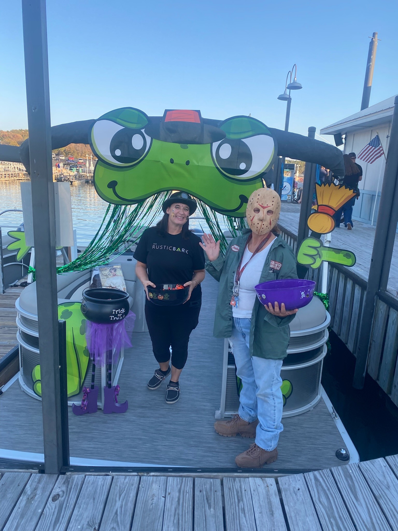 Two people stand on a docked boat with a large, cartoonish frog decoration featuring a witch hat and black fringe hair, surrounded by Halloween-themed props such as a broom and cauldron, under a clear blue sky by the waterfront.