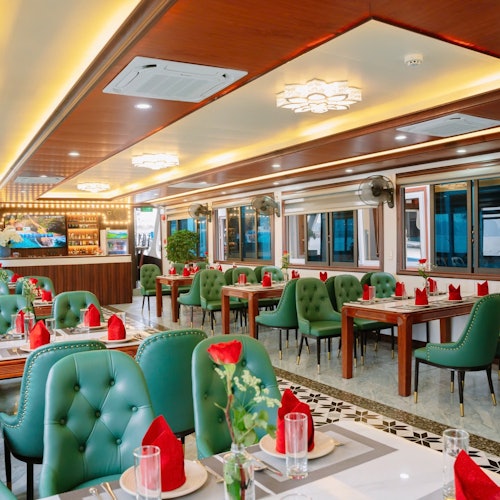 Elegant restaurantinteriør med grønne polstrede stole, træborde dækket med røde servietter og glasvarer, og varm belysning fra lysekroner.