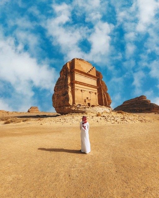 A man in traditional Saudi attire stands before the iconic Al Ula rock-cut tomb under a clear blue sky, representing the rich heritage and legal traditions that SAA Law Firm upholds in Saudi Arabia.