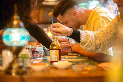 A person crafting a Turkish mosaic lamp, carefully applying multicolored beads to the ornament. The background shows a workshop setting with tools hanging on the wall.