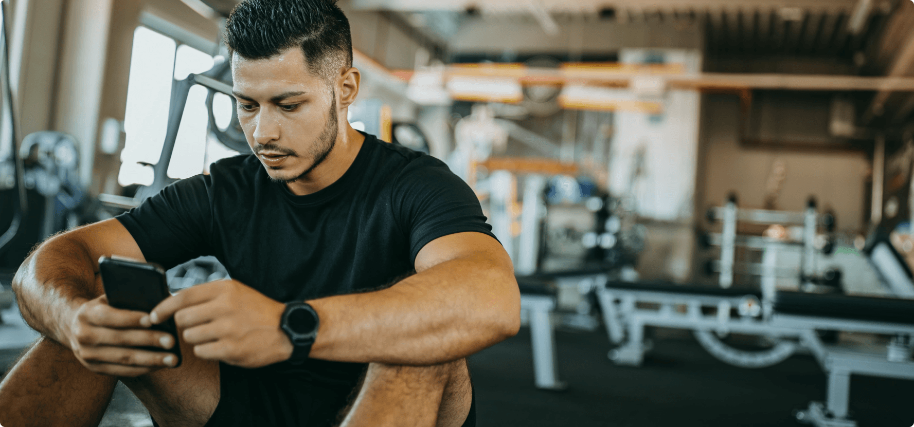 A man in the gym sits in a relaxed position, focused on his phone