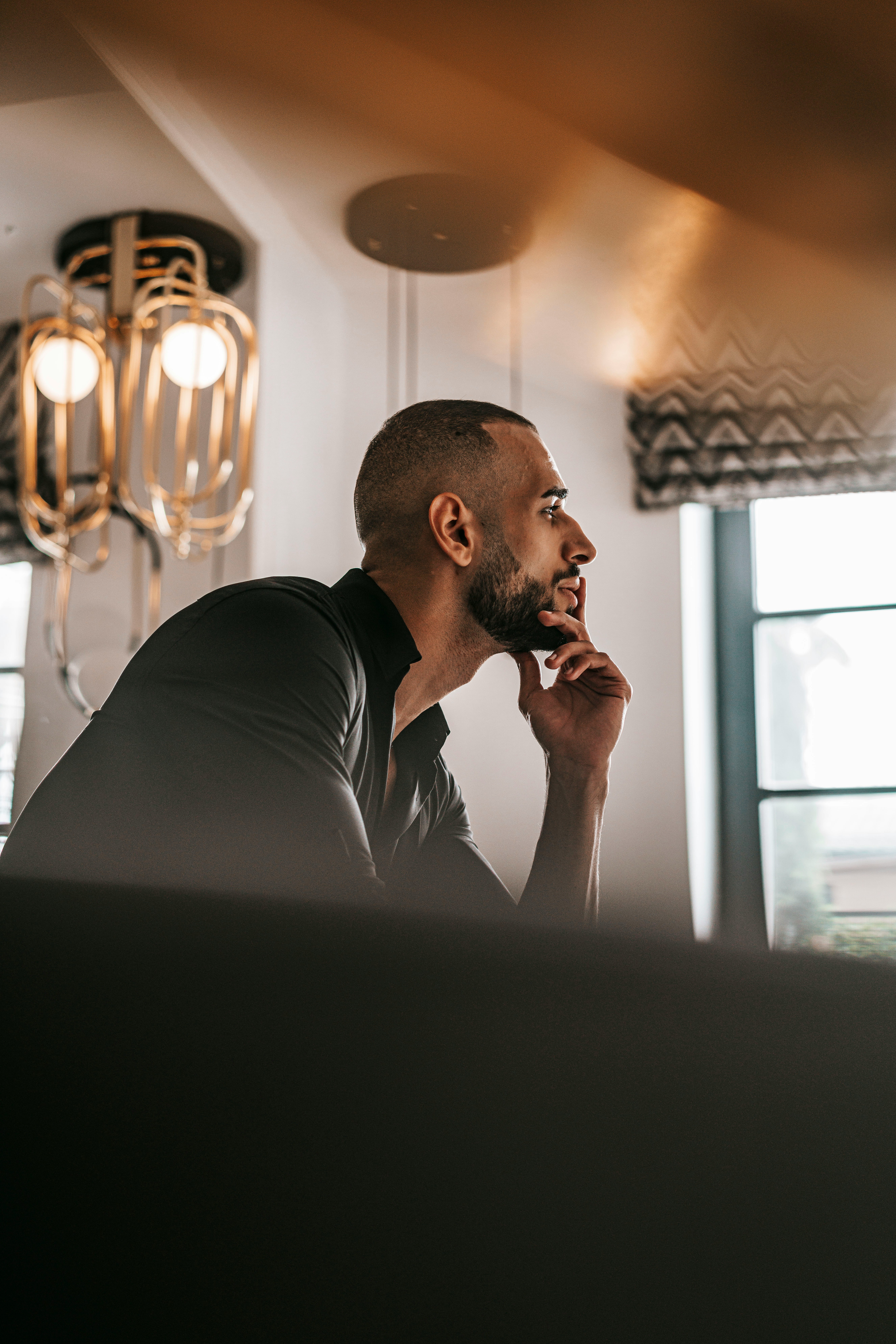 A person deep in thought, resting their chin on their hand, in a well-lit room with decorative lighting.