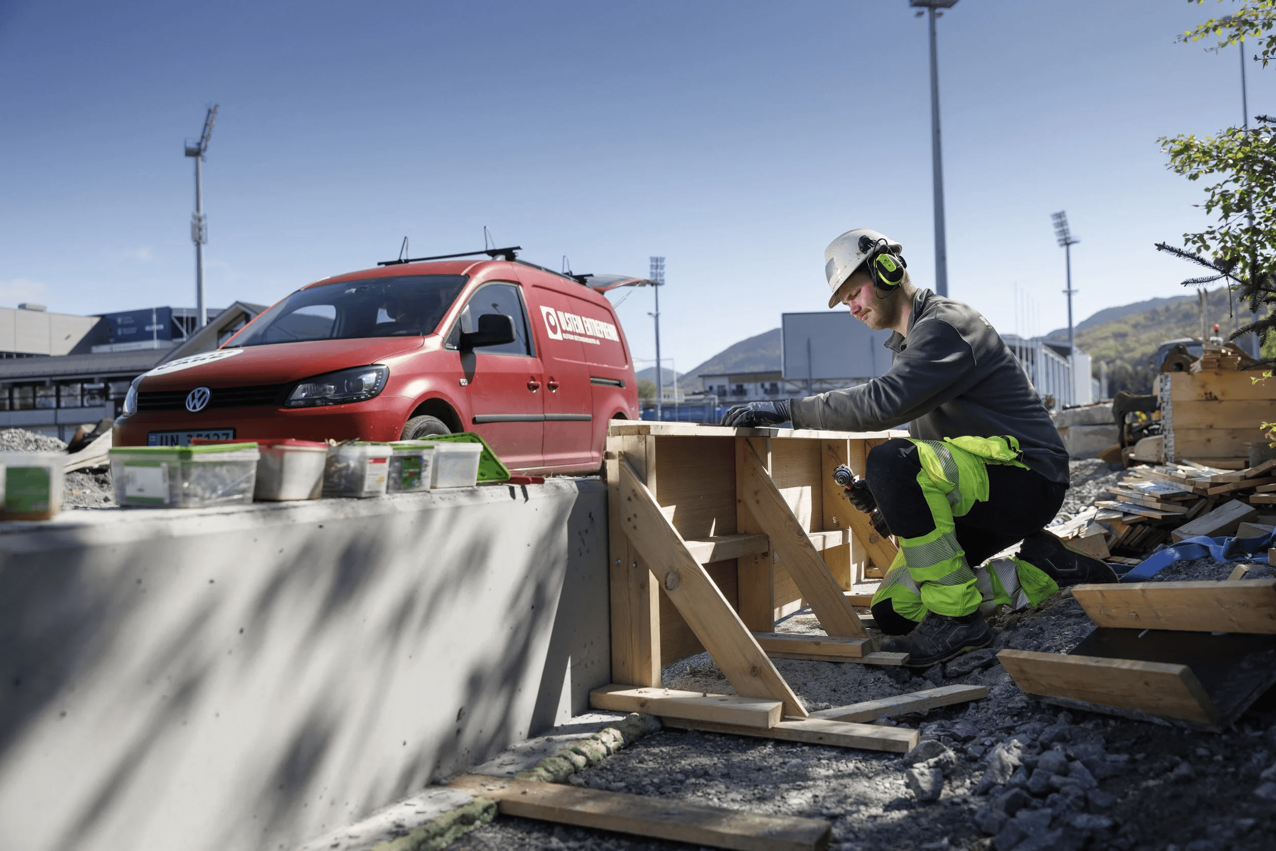 A person sits on a snowy slope near a red van, surrounded by ski equipment and scenic mountains in the background.