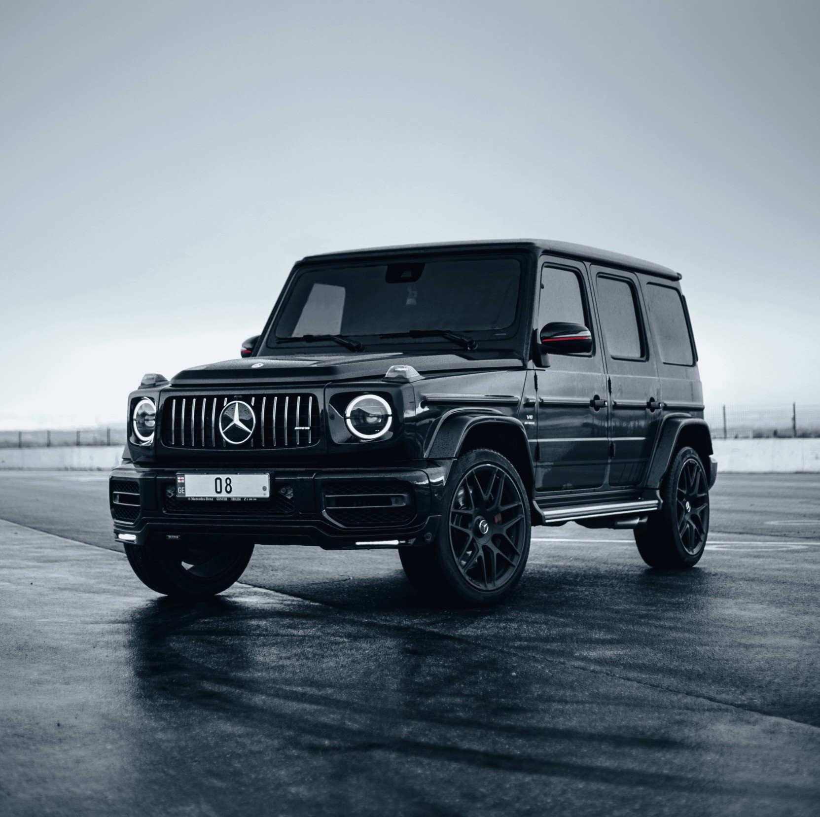 A sleek, black Mercedes-Benz G-Class SUV is parked on a wet road with an overcast sky in the background, showcasing its bold design and luxury features.