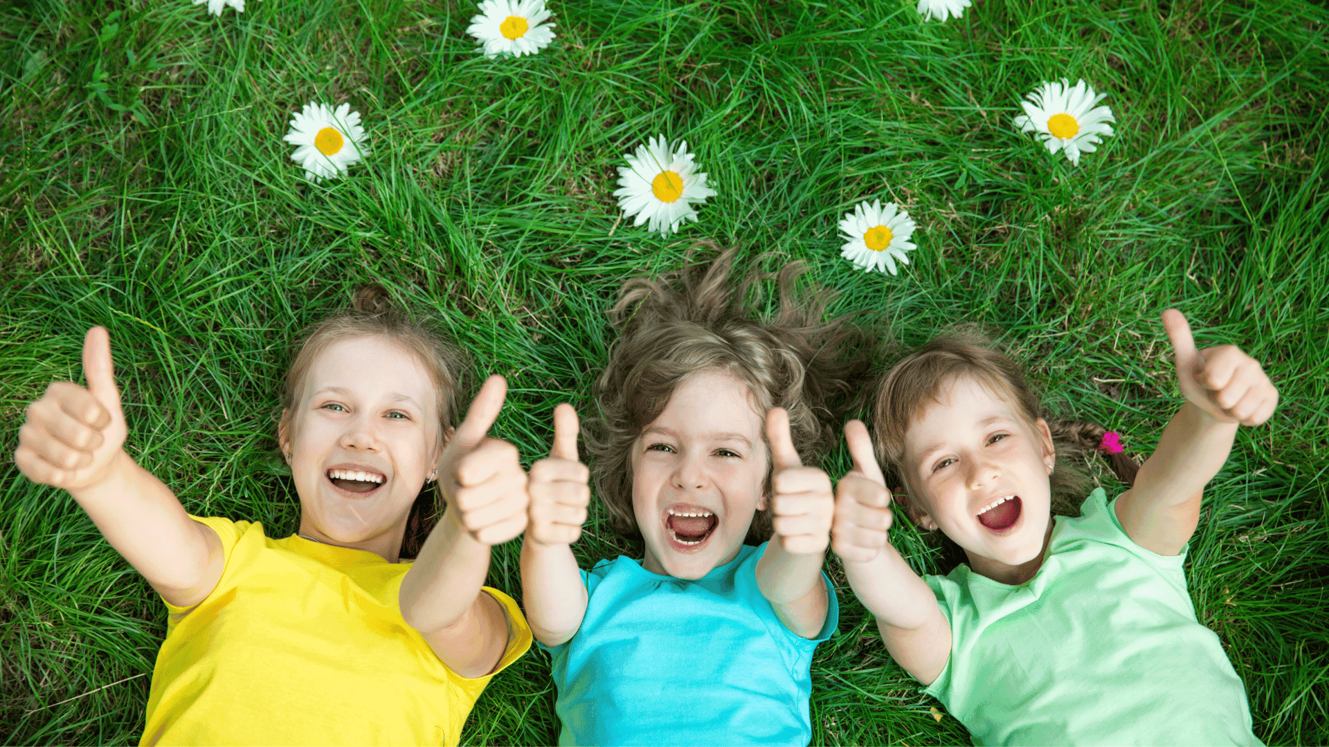 Three children lying on the grass surrounded by flowers, all giving a thumbs up and smiling.