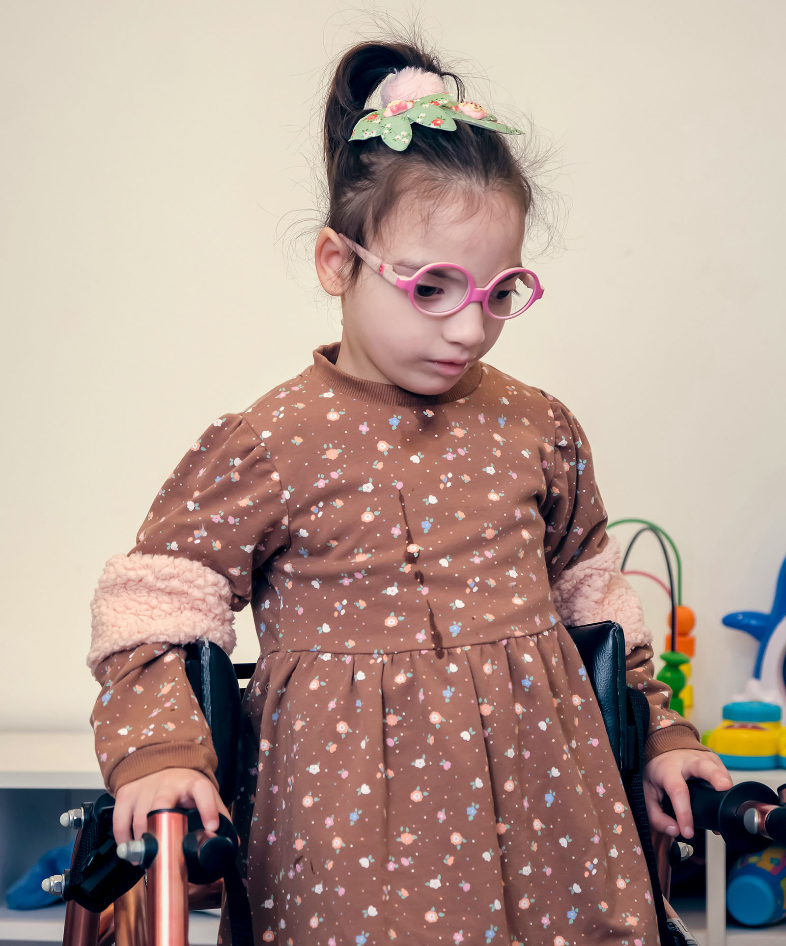 A young girl in a brown dress and pink glasses uses a walker.