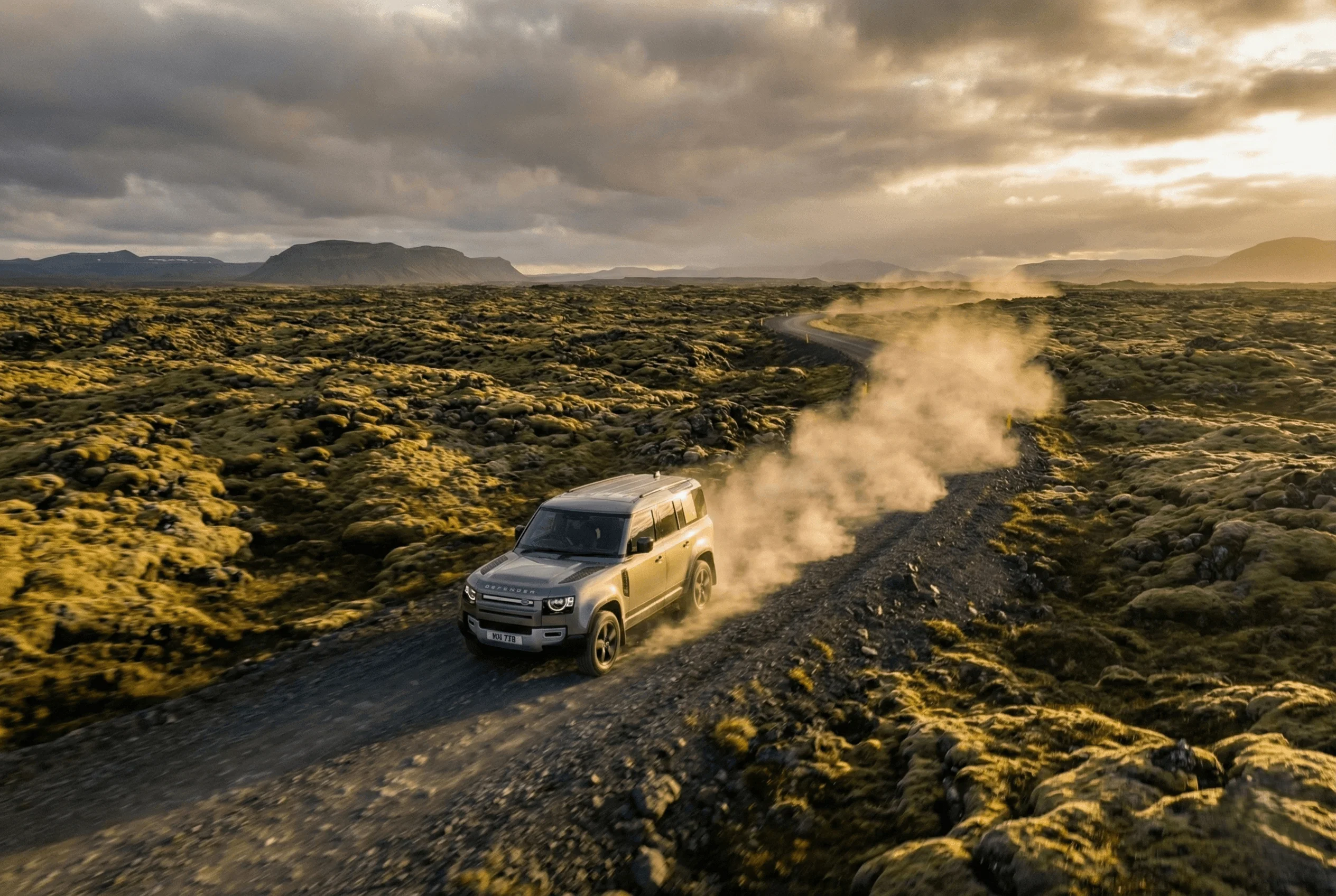 High-angle view of a grey Land Rover Defender driving on a gravel road through a mossy lava field at golden hour.