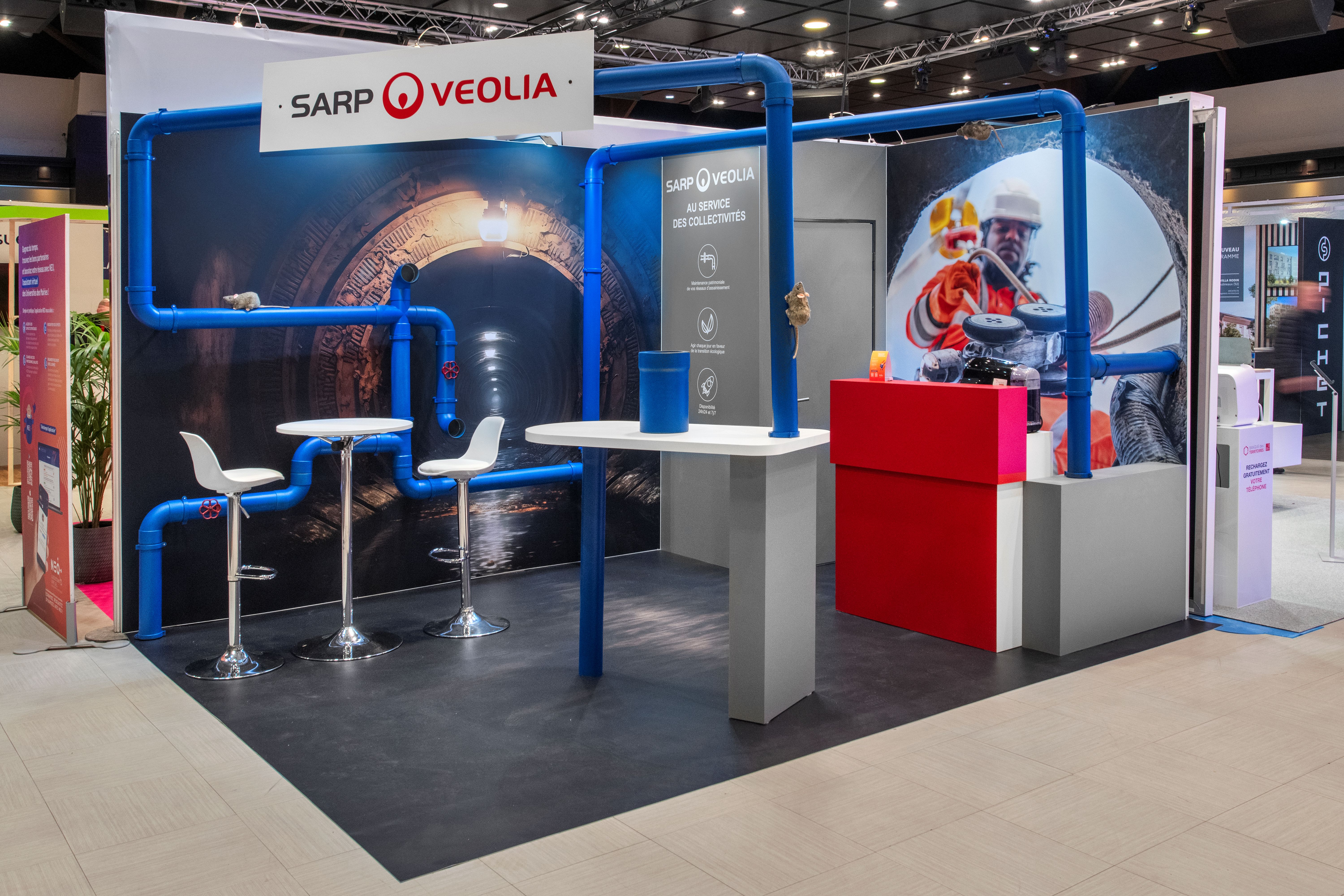Stand SARP Veolia salon UMOP 2025 réalisé par Expace – vue frontale avec structure de canalisations bleues, visuels grand format tunnel et technicien assainissement, totem de présentation et espace lounge