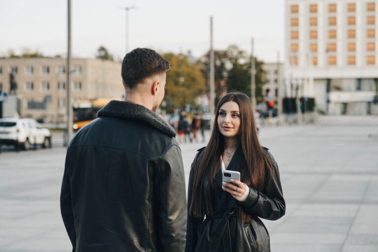 eine brünette frau sieht lasse landeck an, sie stehen beide auf einem platz in einer stadt