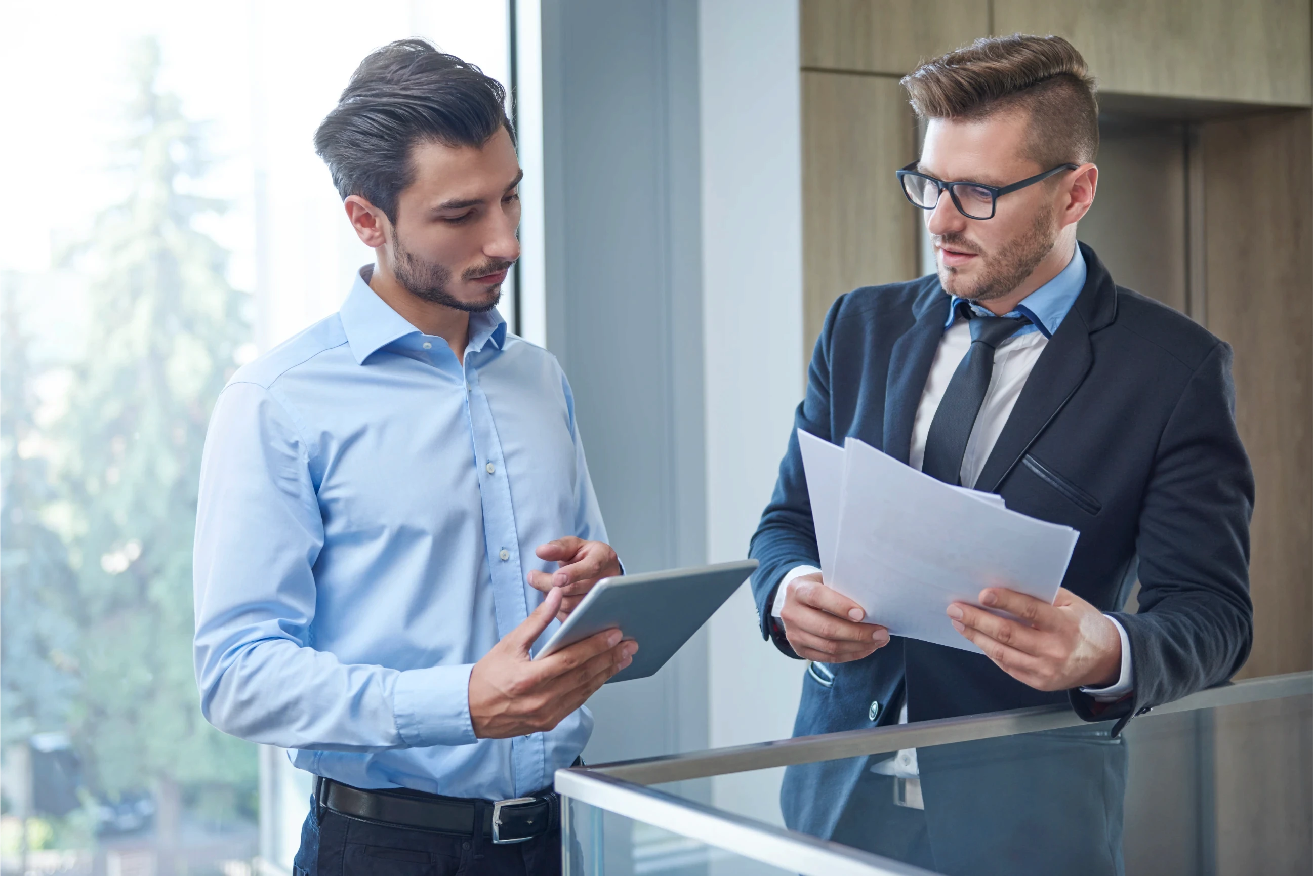 Two men in a modern office engage in a discussion. One, holding a tablet, listens attentively; the other, in a suit, explains while holding papers.