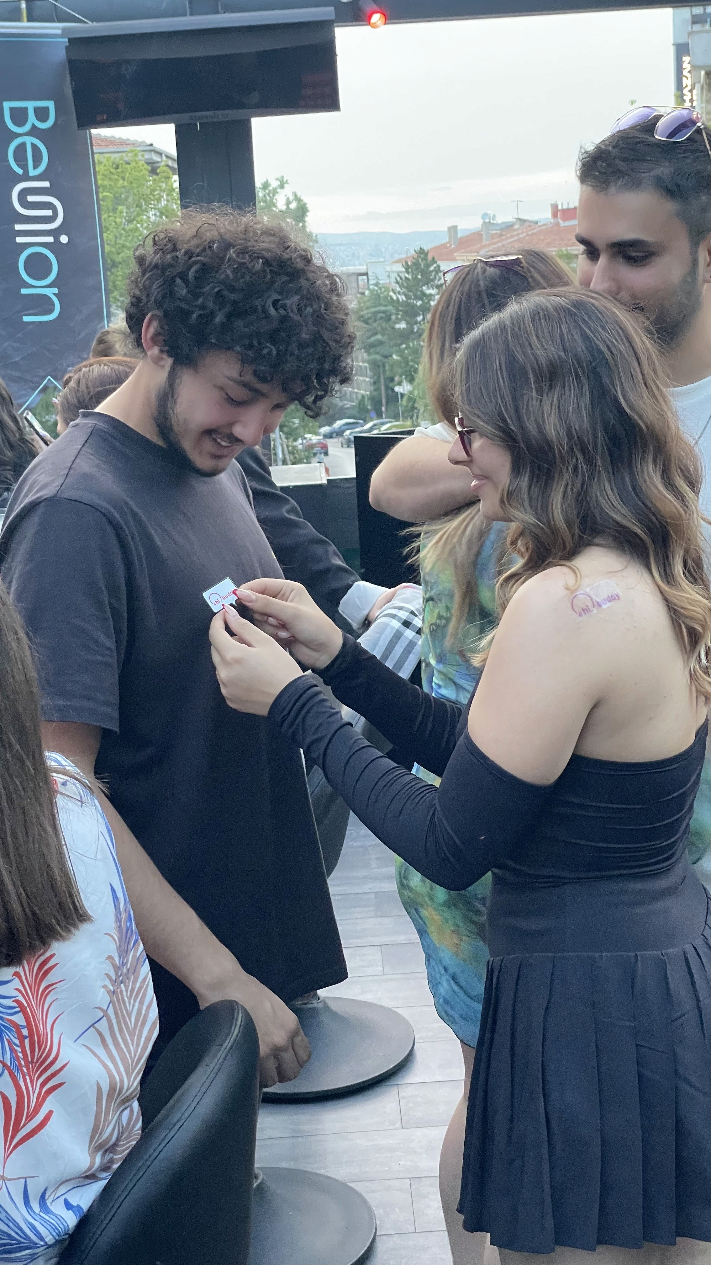 A woman places a 'hibuddy' sticker on her new friend, smiling as they bond during a fun social activity.