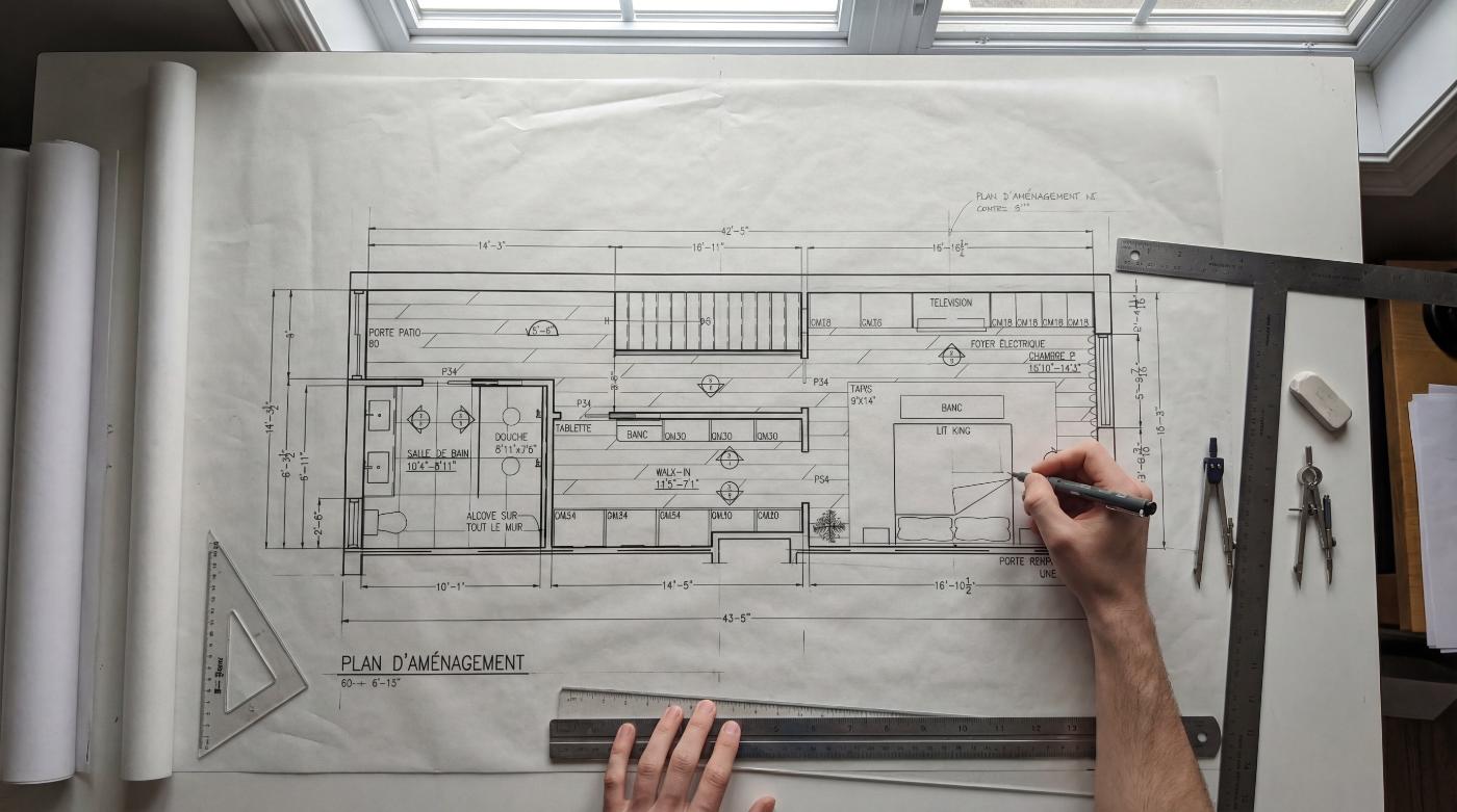 Plan d'aménagement posé sur un bureau. Deux mains sont visibles, celle en bas de l'image tient une règle, tandis que celle de droite tient un crayon. En haut, On aperçoit le bas de la fenêtre.