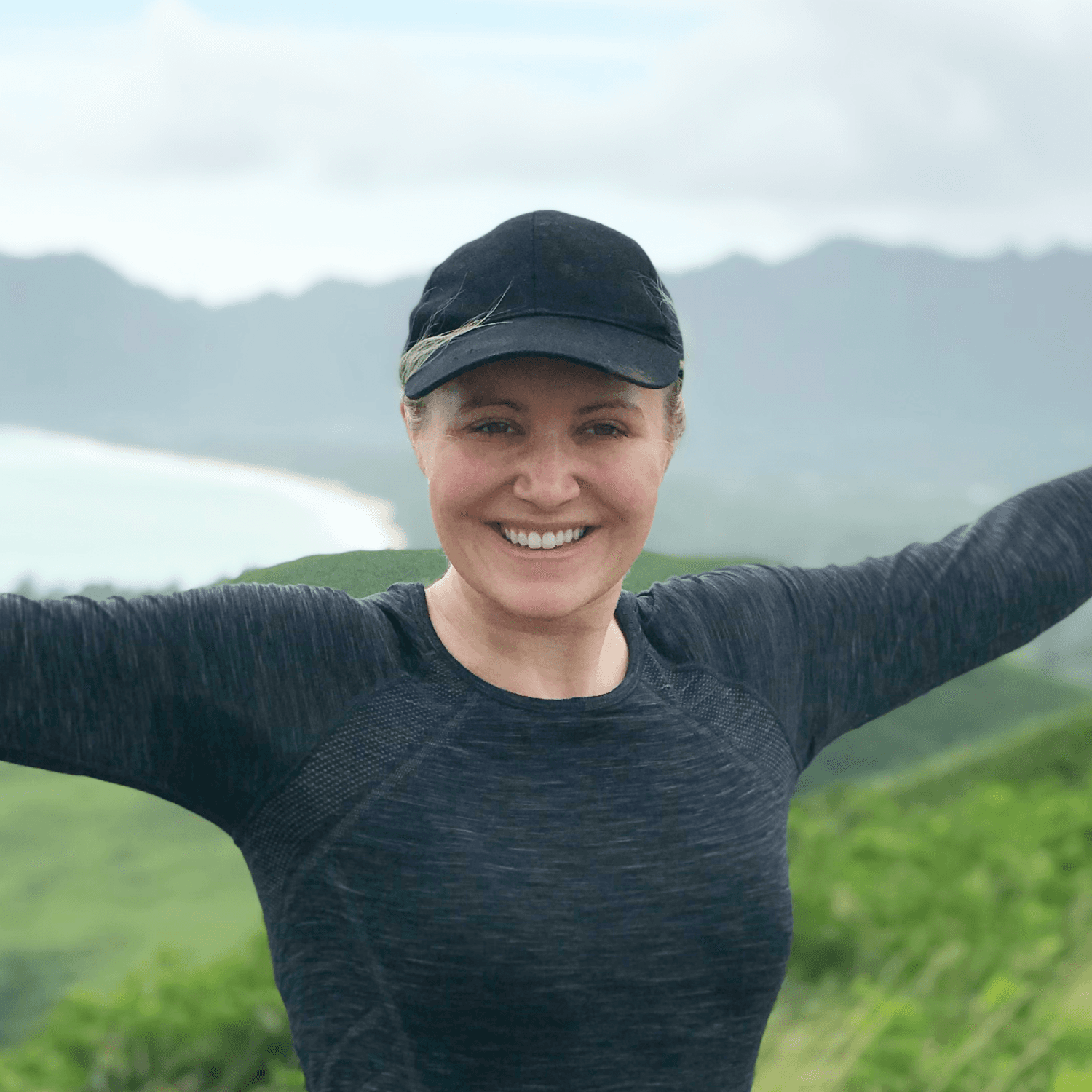 Bec Martin stands outdoors on a grassy hill, smiling with arms outstretched, with mountains and a bay in the background.