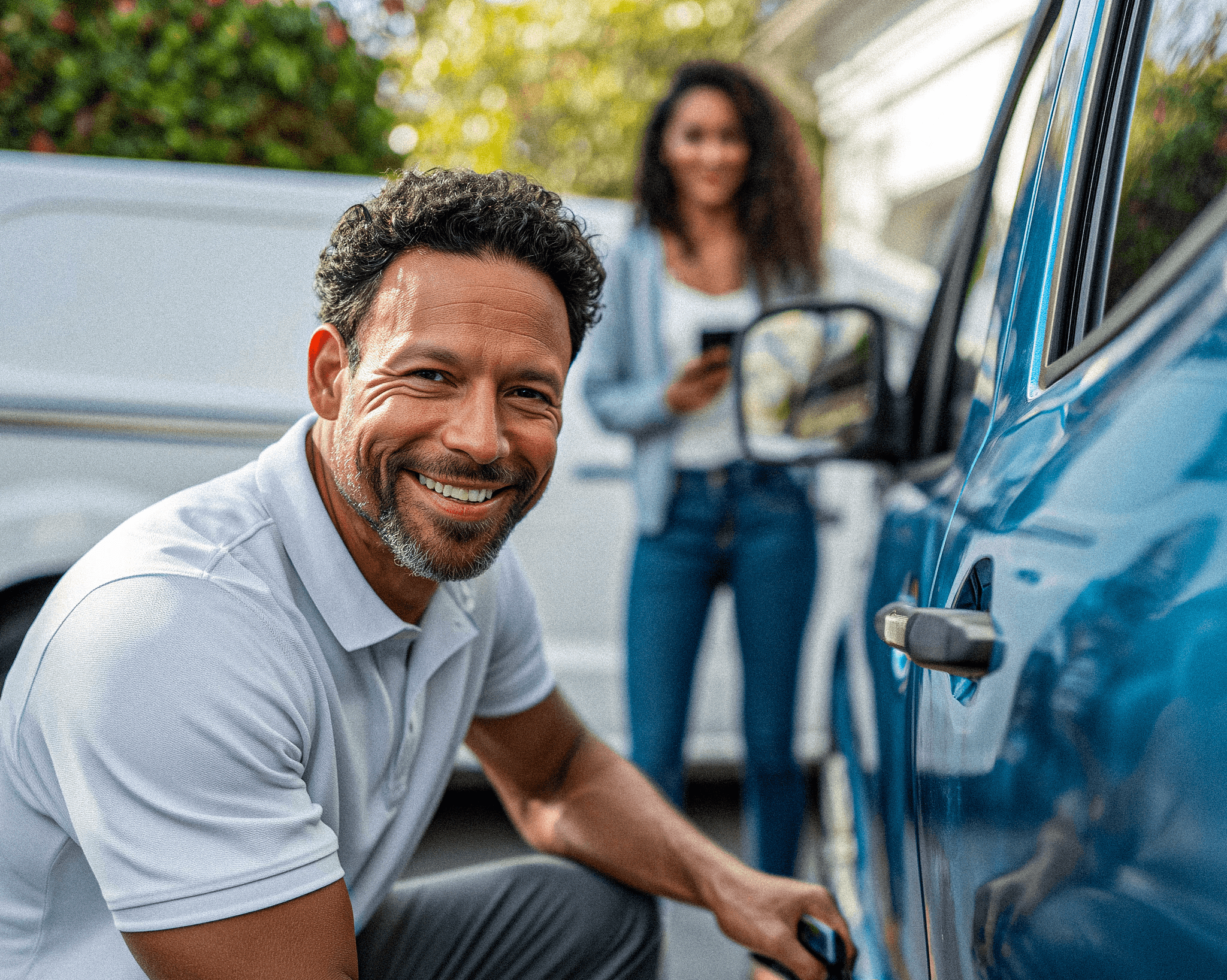 A locksmith man in Minneapolis in a white polo shirt kneels beside a blue car, smiling while unlocking the car door, with a woman in the background holding a smartphone, both outdoors on a sunny day. Suggesting a professional auto locksmith service.