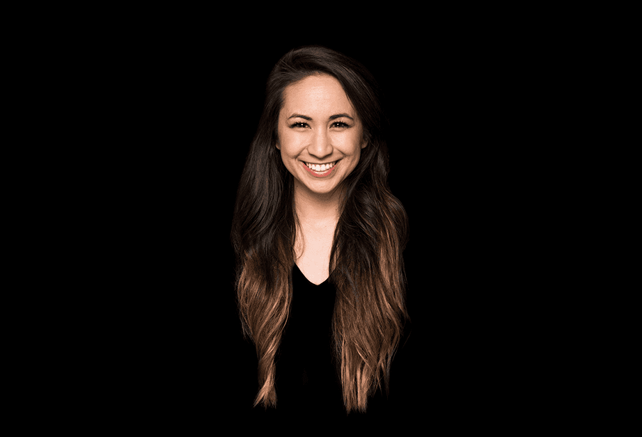A woman with long brown hair and a black top smiles at the camera against a solid black background. Her hair falls around her shoulders and frames her face. The lighting highlights her features and hair texture.
