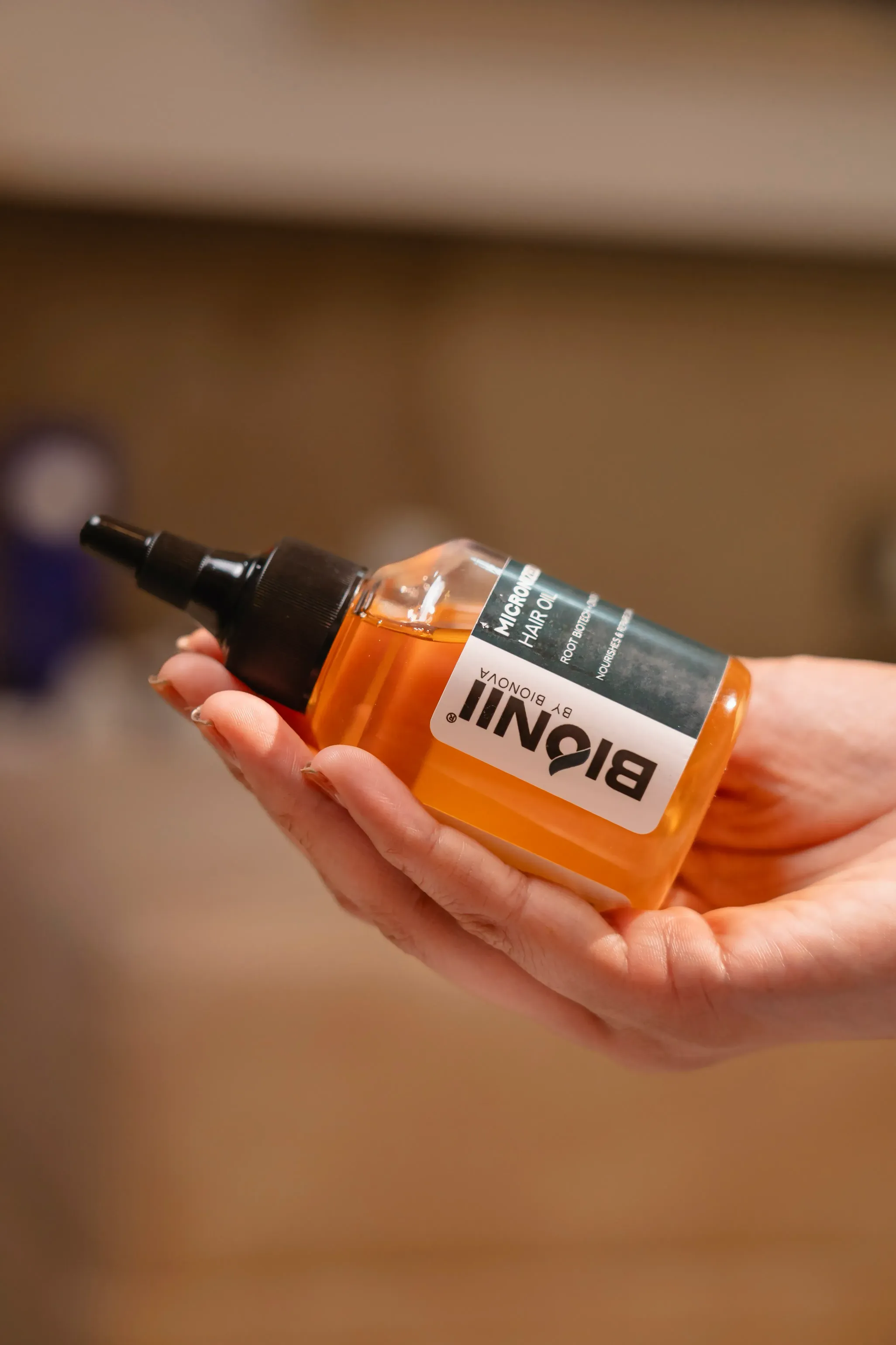 A hand holding a bottle of liquid with a dropper top, set against a blurred background.