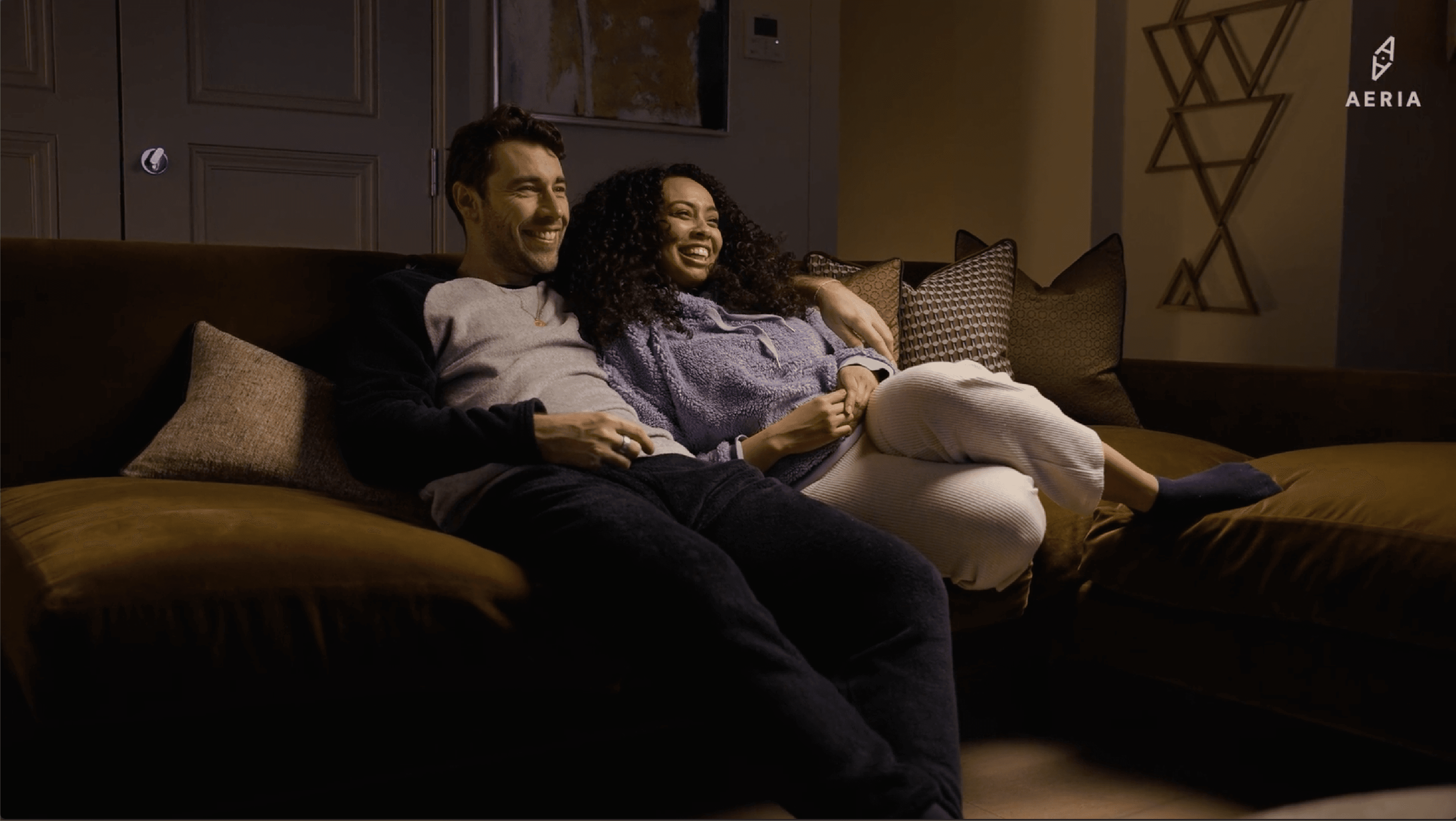 A couple sitting close together on a dark sofa in a warmly lit living room, smiling and relaxing with their arms around each other. Decorative cushions and wall art are visible in the background, and a logo reading “AERIA” appears in the top right corner.