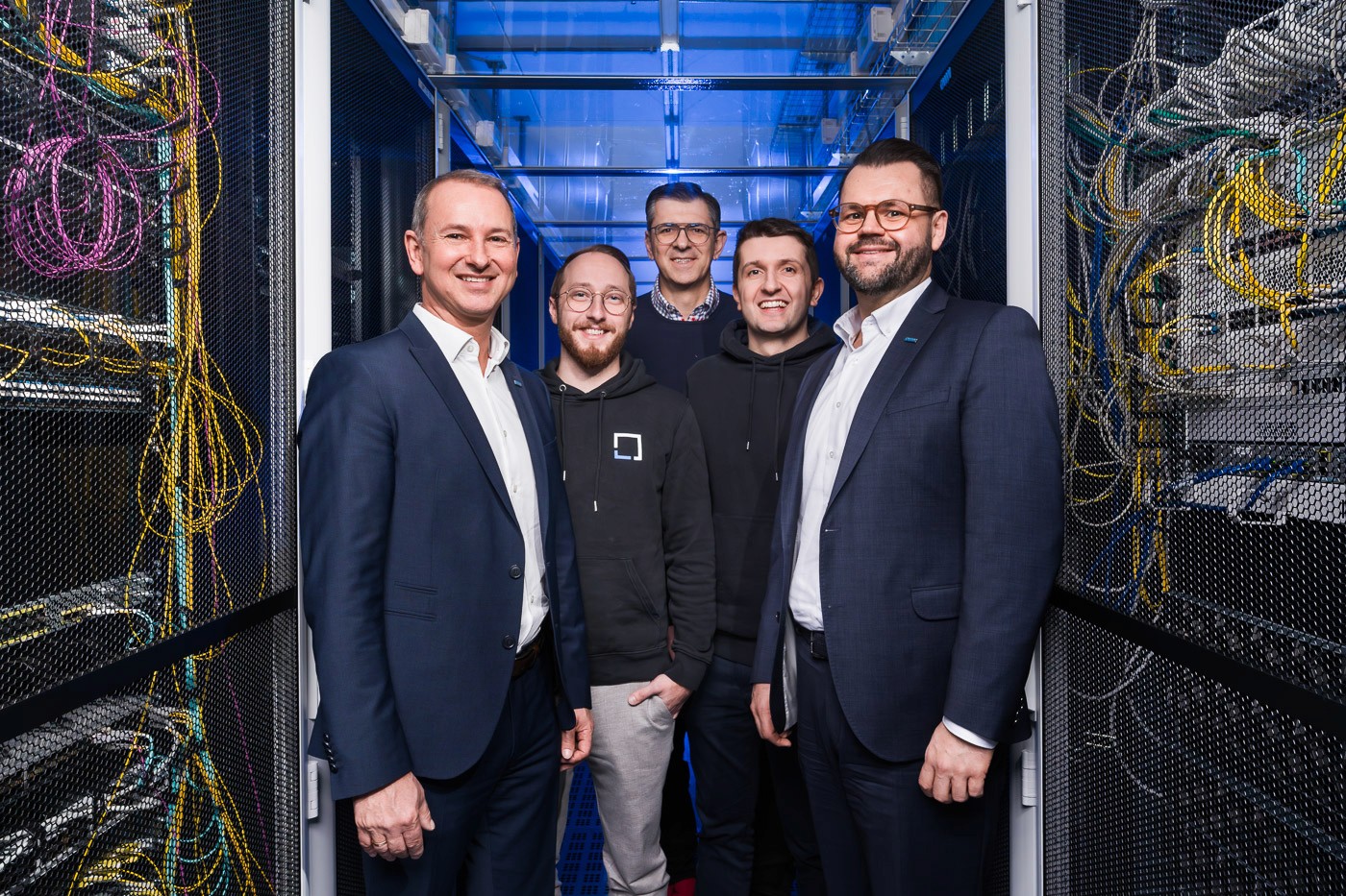 Fünf Männer, die nebeneinander zwischen Serverracks stehen.