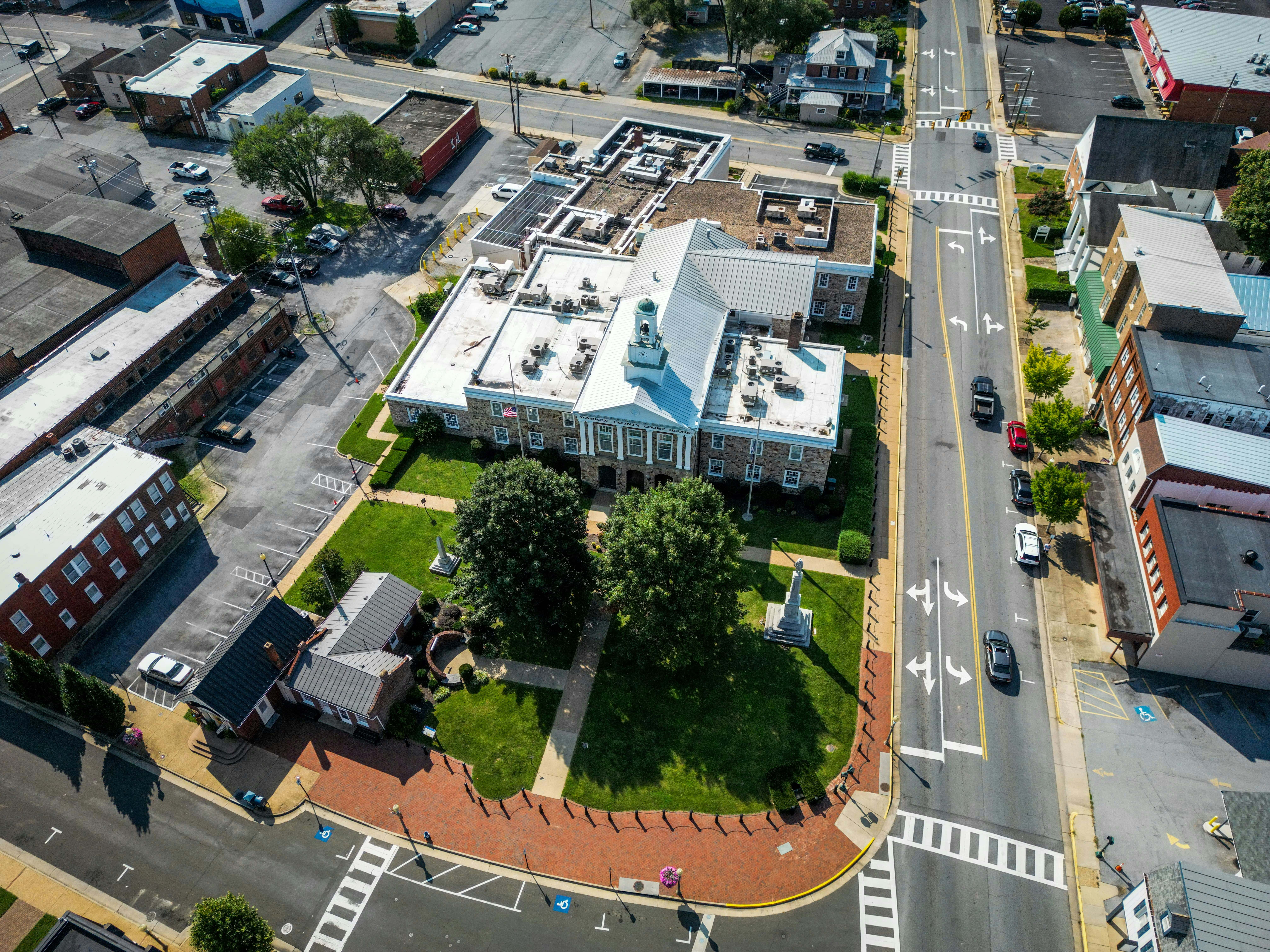 Aerial view of a historic courthouse square in a small Virginia town, surrounded by green lawns, trees, and red-brick pathways. The surrounding area features streets with marked crosswalks, clear traffic lanes, and a mix of commercial and residential buildings, characteristic of Virginia’s traditional town layout.