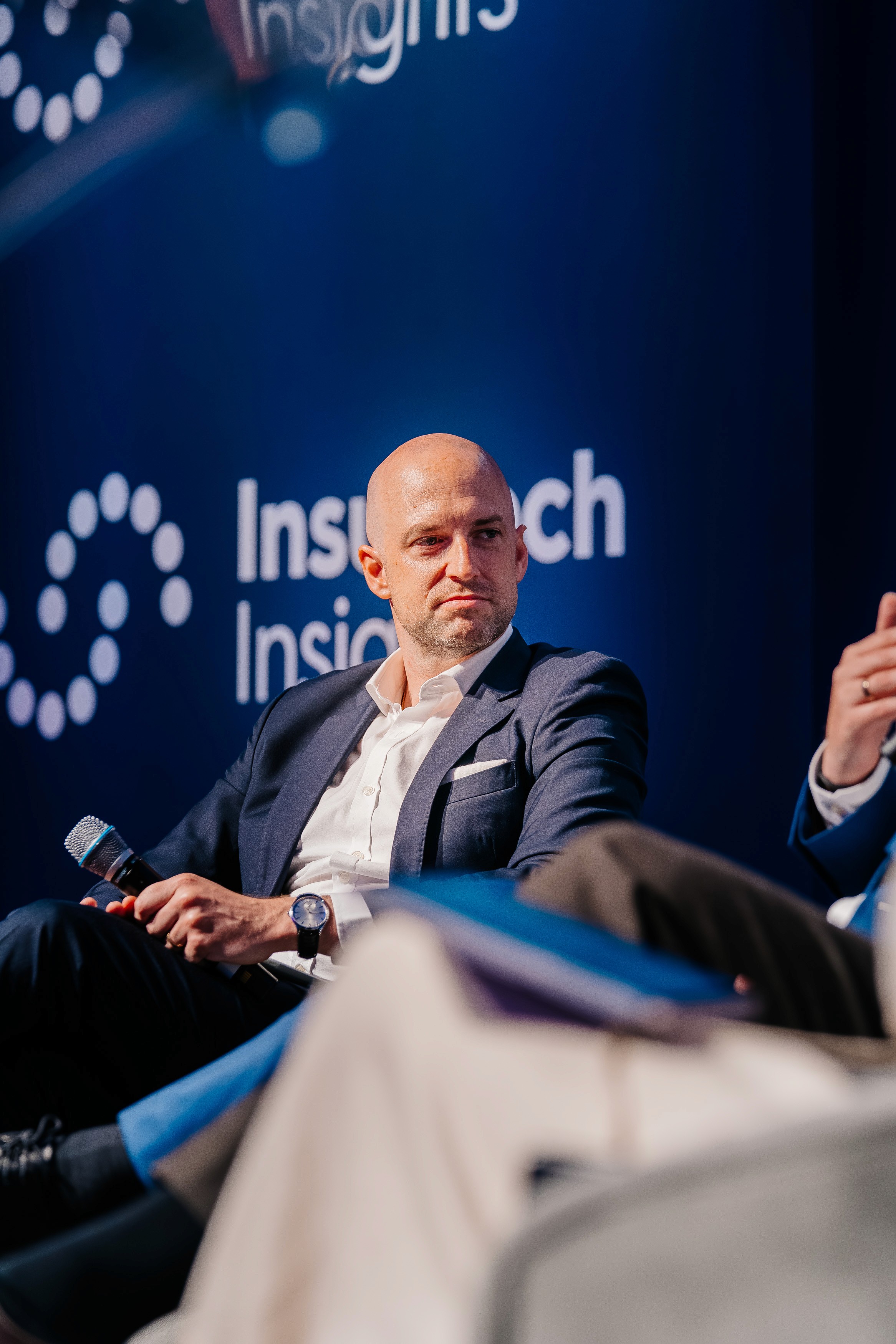 Two individuals engaged in a discussion on stage, with a blue background displaying the logo of a research institution.