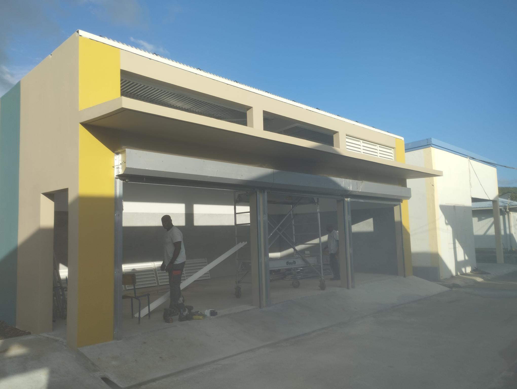 Modern building with yellow accents under a clear blue sky. Two workers visible near an open garage area, tools scattered. Calm, sunny atmosphere.