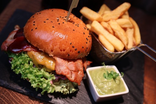 Bacon-Cheeseburger im kAlps mit Sesambrötchen, geschmolzenem Käse, Salat, knusprigem Speck, Pommes und grünem Dip.