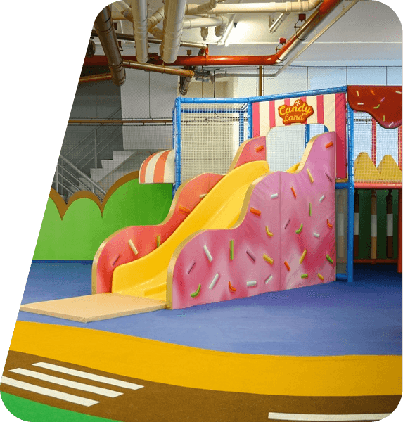 Colorful playground with intricate climbing structures, including a netted white and orange dome, set on a bright, winding path. Lush greenery surrounds the area, giving it an inviting and vibrant atmosphere.