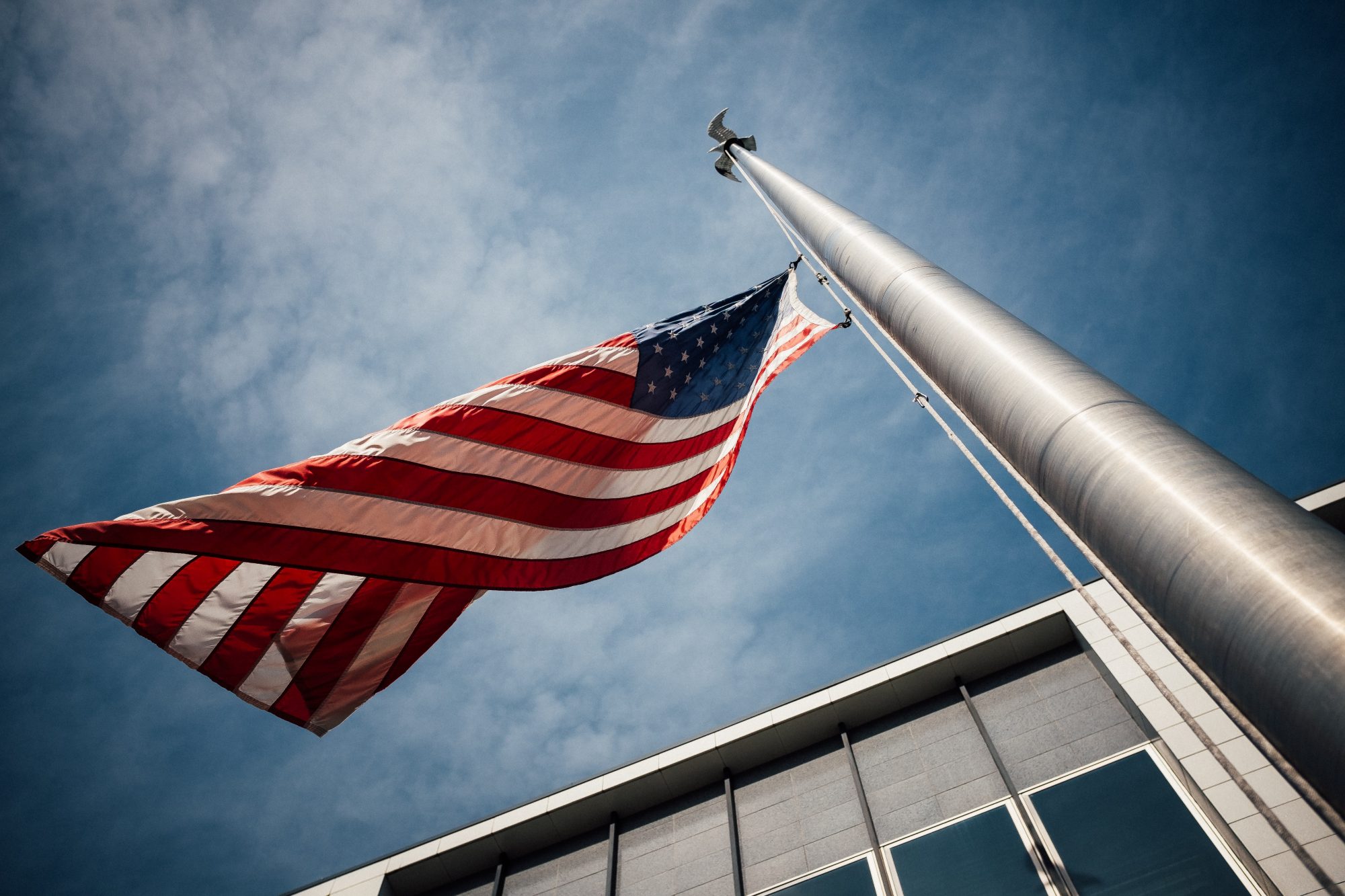 American flag waving on a pole against a blue sky with clouds, in front of a modern building.