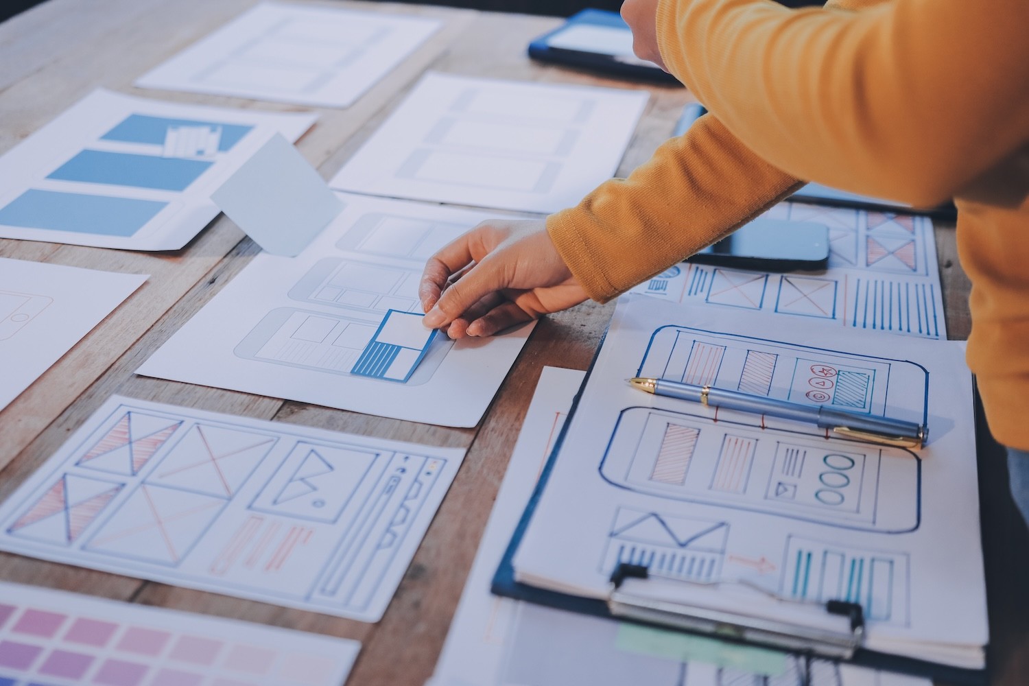 Hand einer Person in orangefarbenem Pullover, die auf einem Tisch mit mehreren Wireframe-Zeichnungen und einem Stift arbeitet