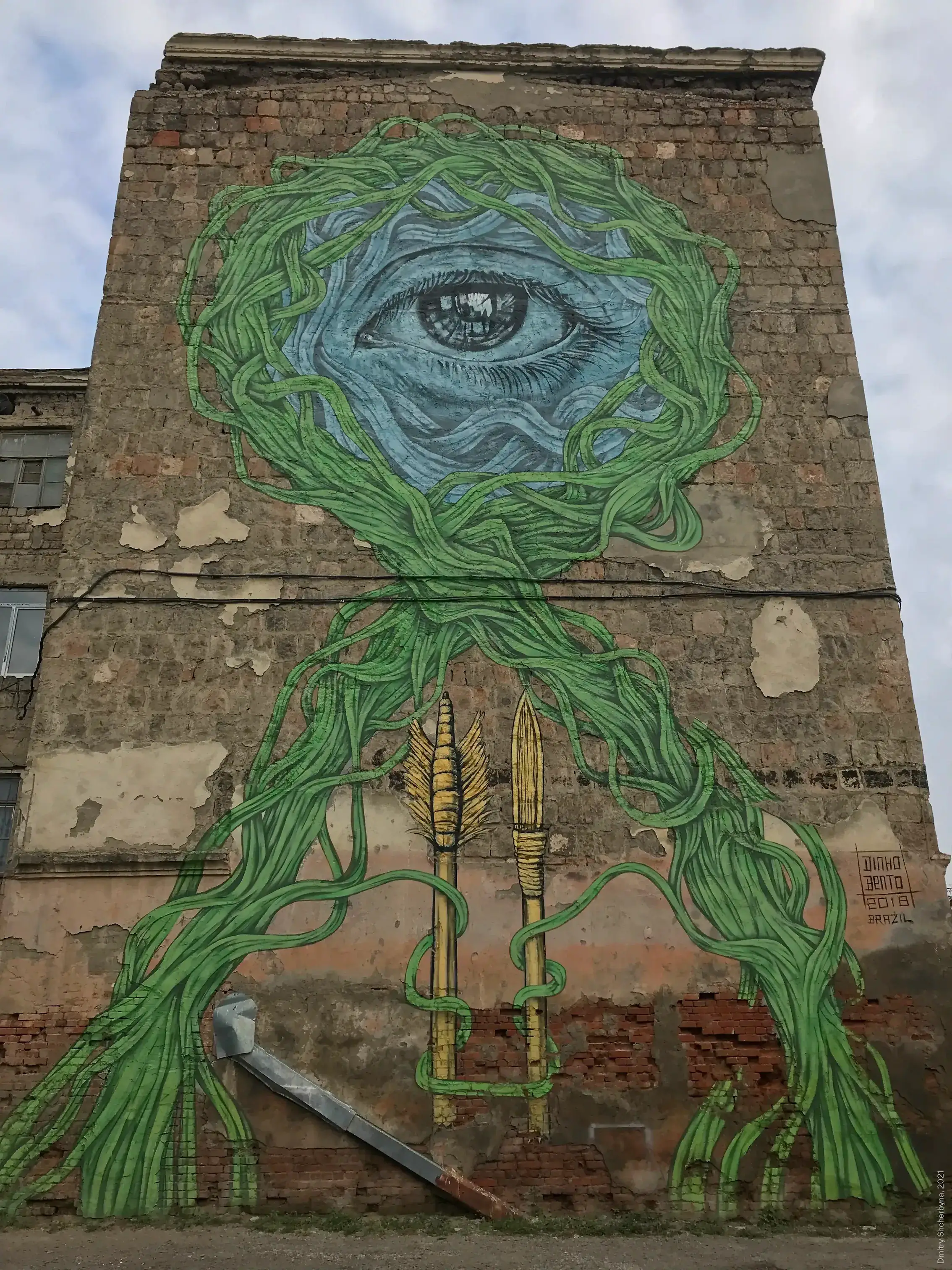 A mural on an exposed brick wall features a large realistic blue eye surrounded by tangled green vines that cascade down around two golden arrow-shaped forms.