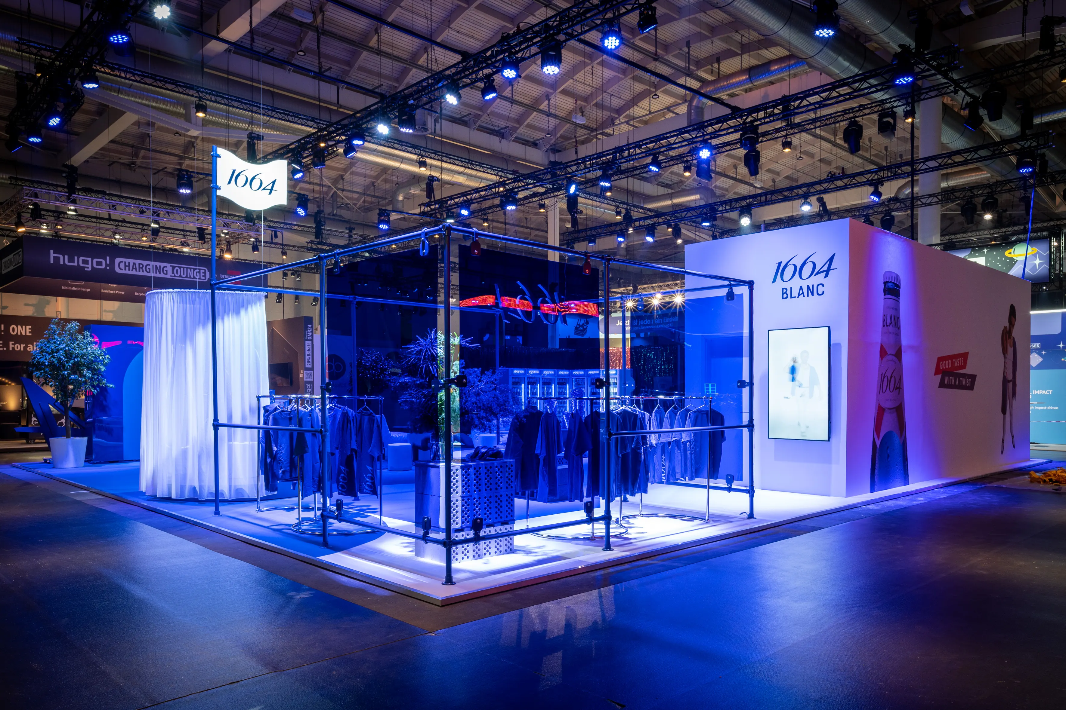 Blue scaffold cube with apparel racks and white curtain at the 1664 Blanc booth under dark trade fair trusses.