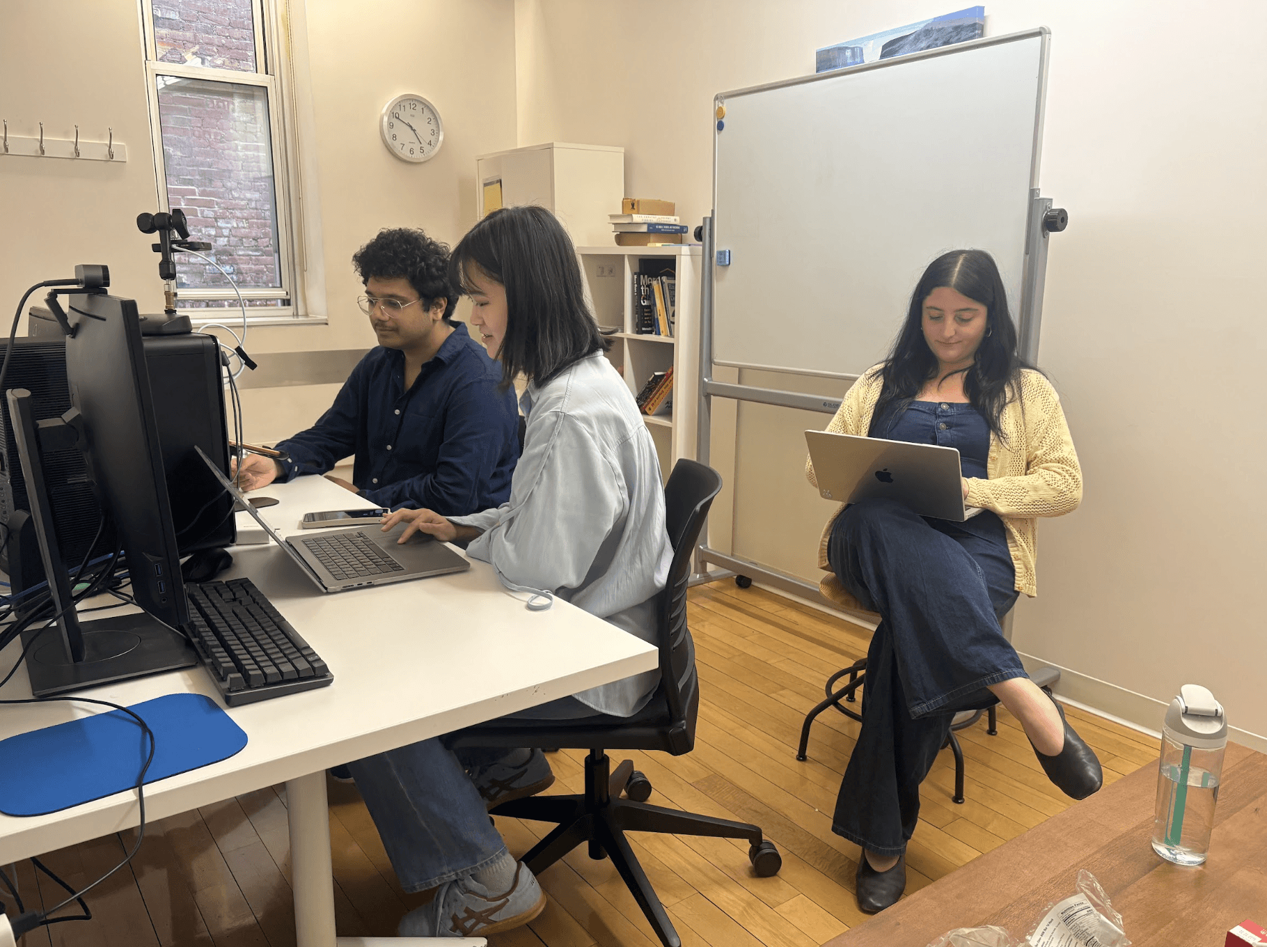 Two team members working in the Pratt UX lab. Two sit at a desk with laptops and recording equipment; the user sits to the side with a laptop, in front of a blank whiteboard.