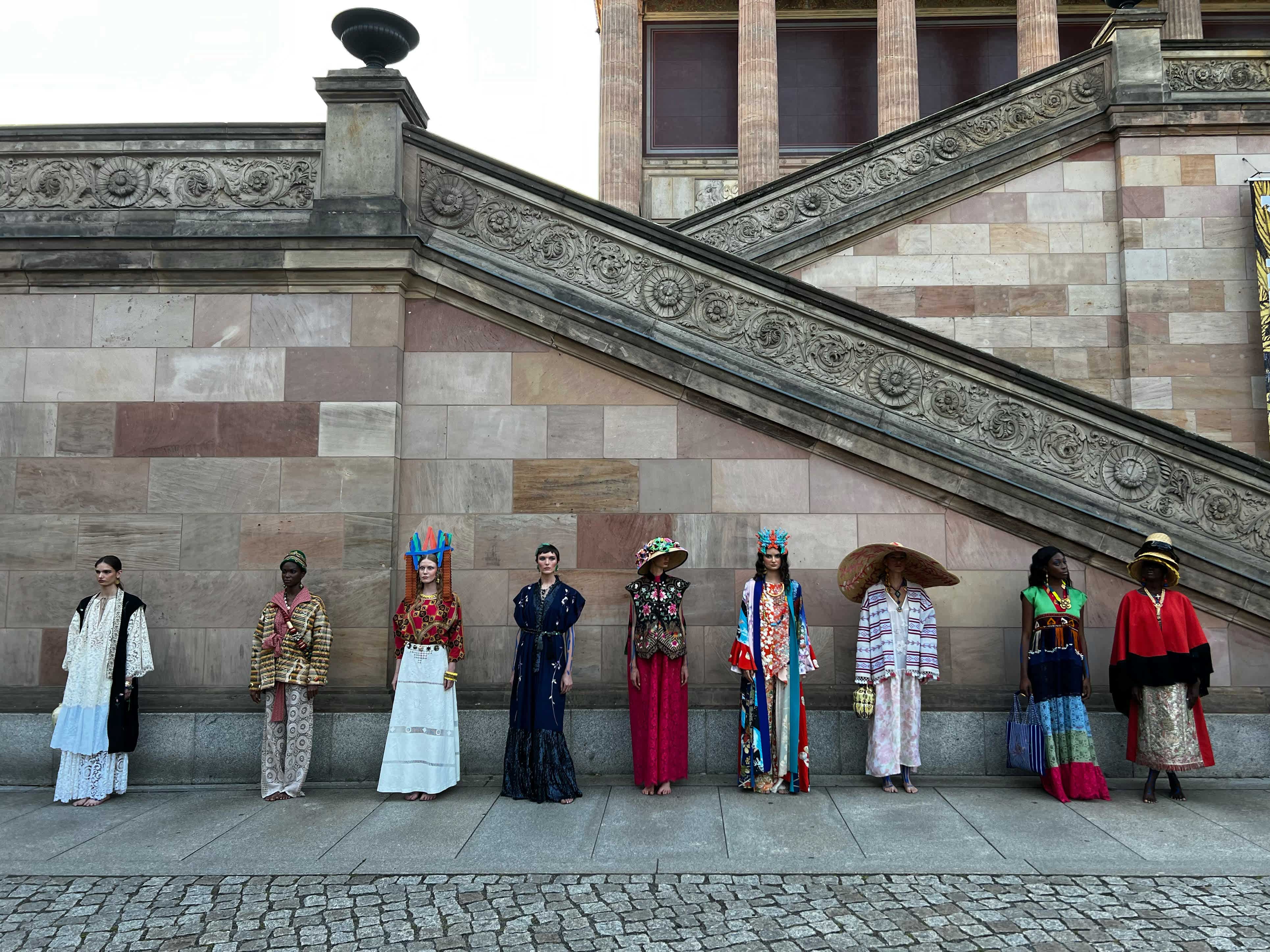 A line of nine fashion models standing side-by-side at the bottom of a wide stone staircase, showcasing various colorful and bohemian outfits from the Rianna + Nina collection.